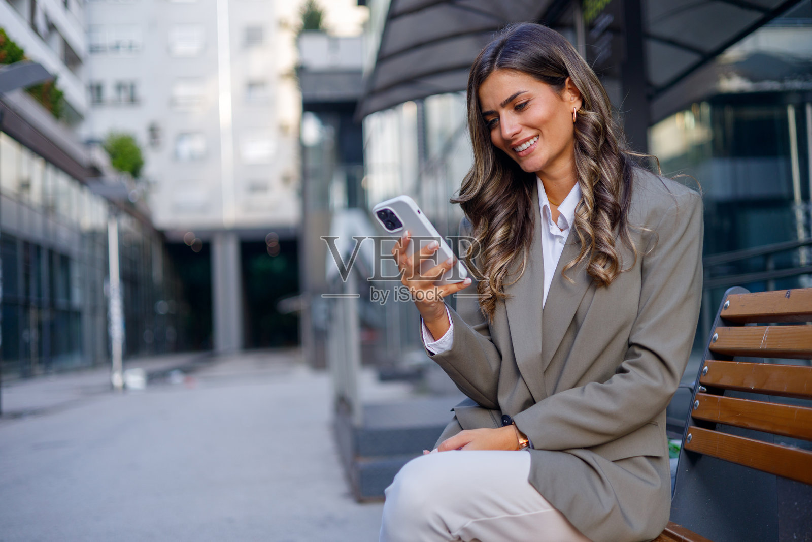 年轻女商人在办公大楼前的长椅上坐着,使用智能手机浏览社交媒体并在线连接。照片摄影图片