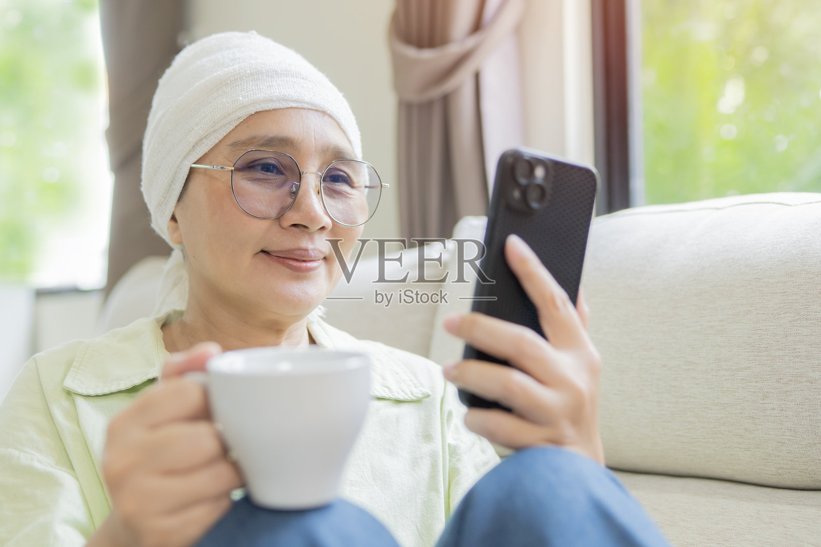 身穿白色围巾的成年女性癌症患者,一边喝茶一边使用手机照片摄影图片