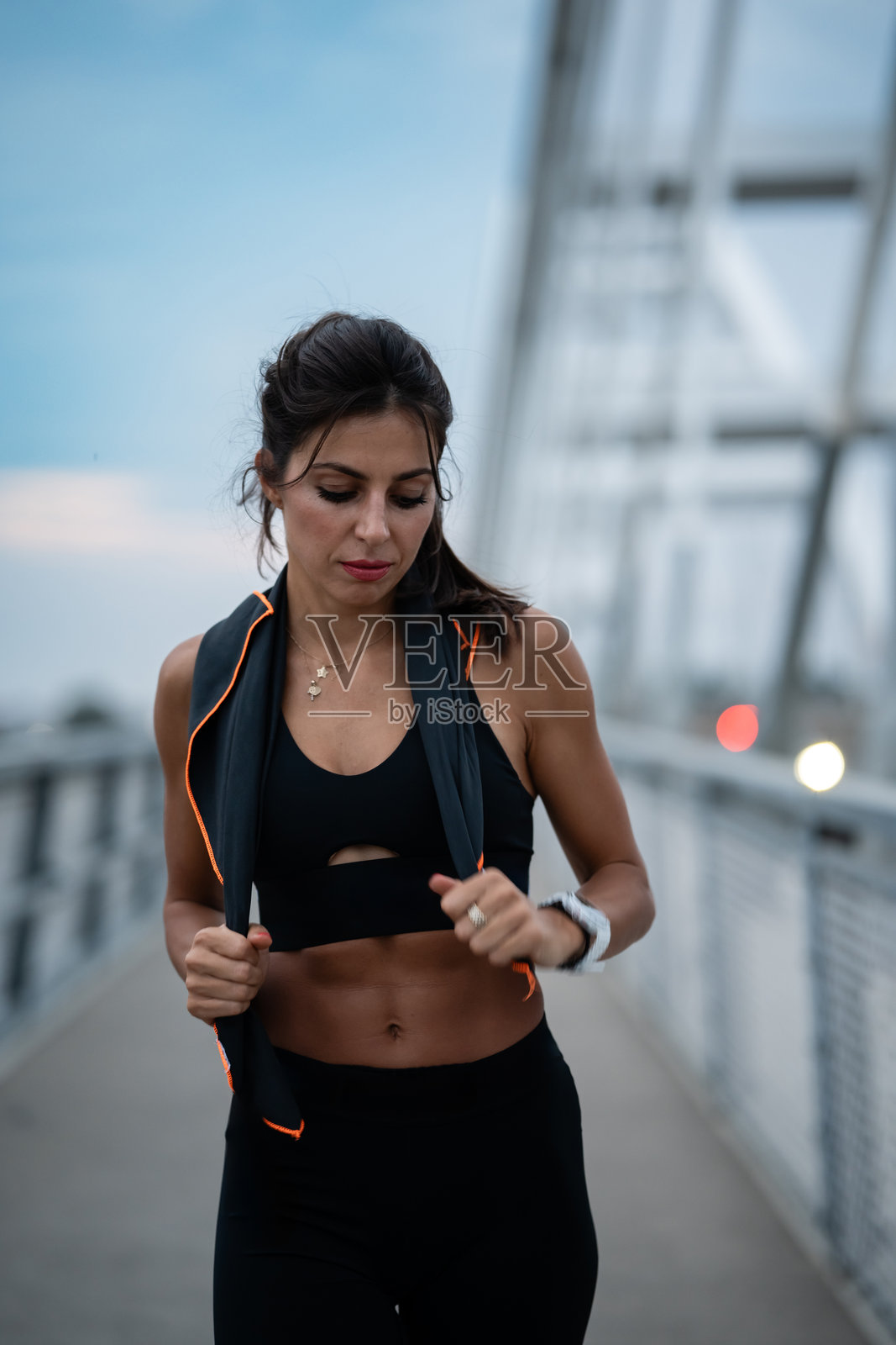 身材健美的女性在日落时分的桥上慢跑,享受城市锻炼并在智能手表上监测进度照片摄影图片