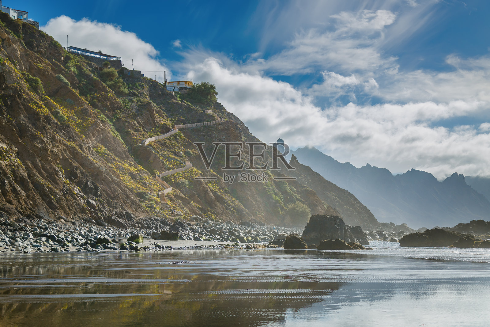 特内里费岛的贝尼霍海滩崎岖的海岸风景,陡峭的岩石山坡上有一条蜿蜒的小路通向建筑,远处是高耸的山峰和在反射海面上动态的天空。照片摄影图片