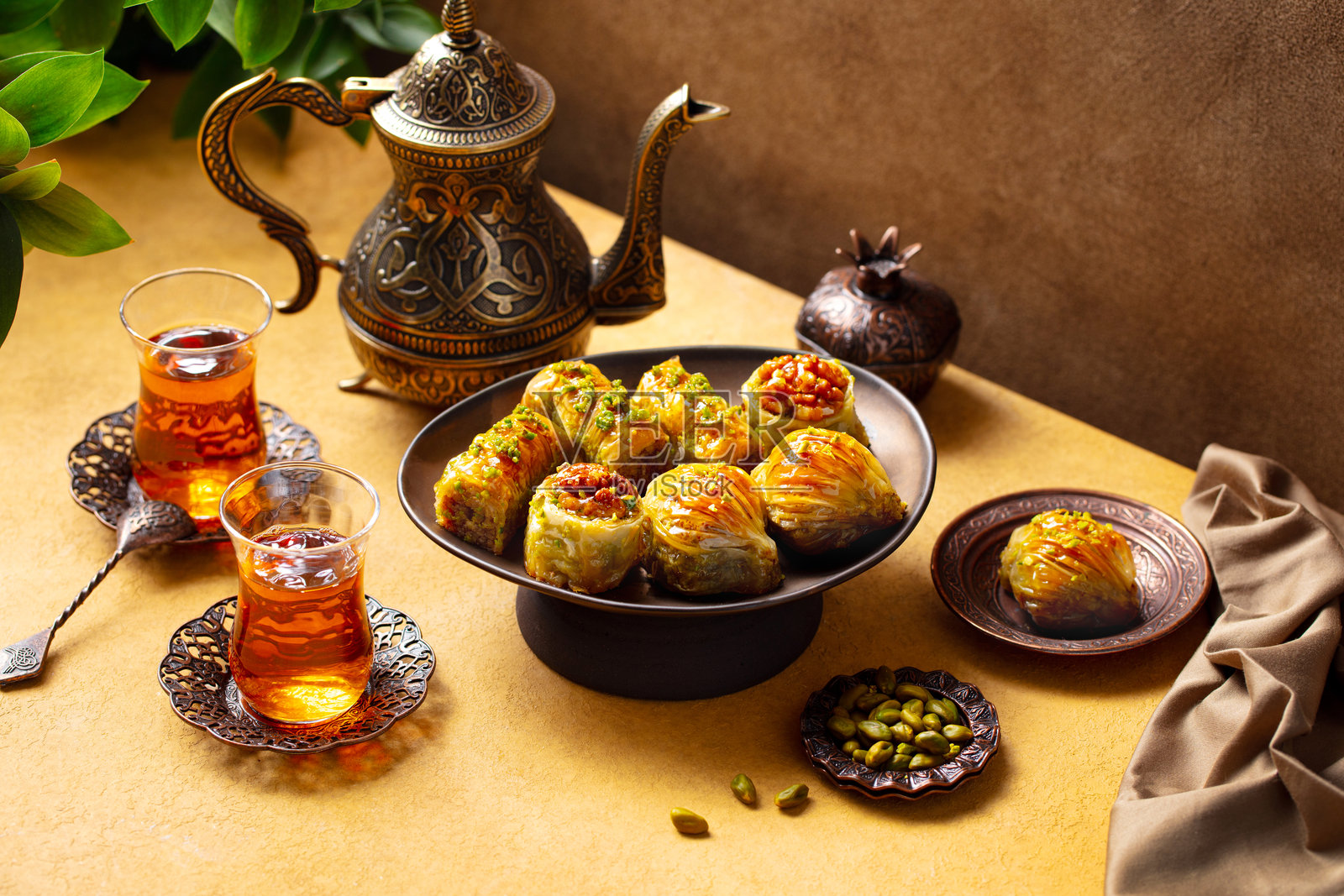 铜茶壶中盛着的阿尔穆杜玻璃茶杯旁的巴克拉瓦甜点,背景为黄色和棕色,特写。照片摄影图片