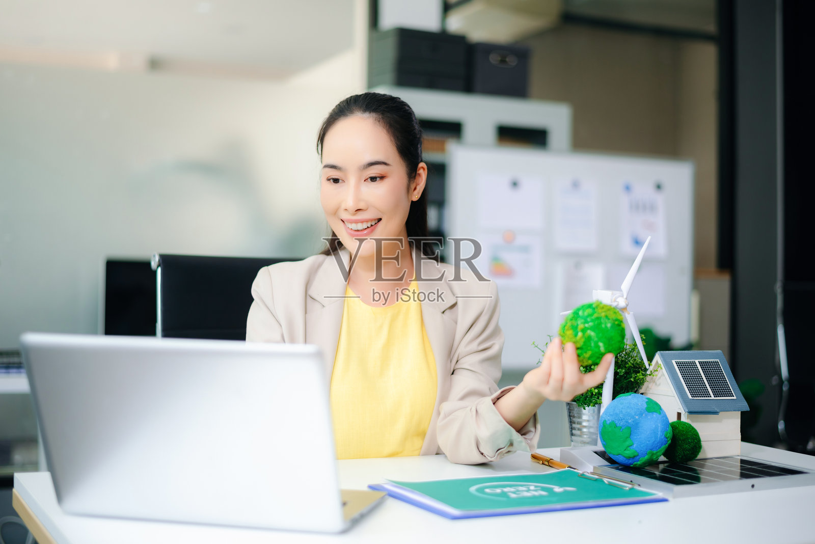 亚洲女性在使用笔记本电脑,同时手握绿色地球仪。周围有风力发电机、太阳能电池板和生态住宅模型。清洁能源和可持续发展的概念。照片摄影图片