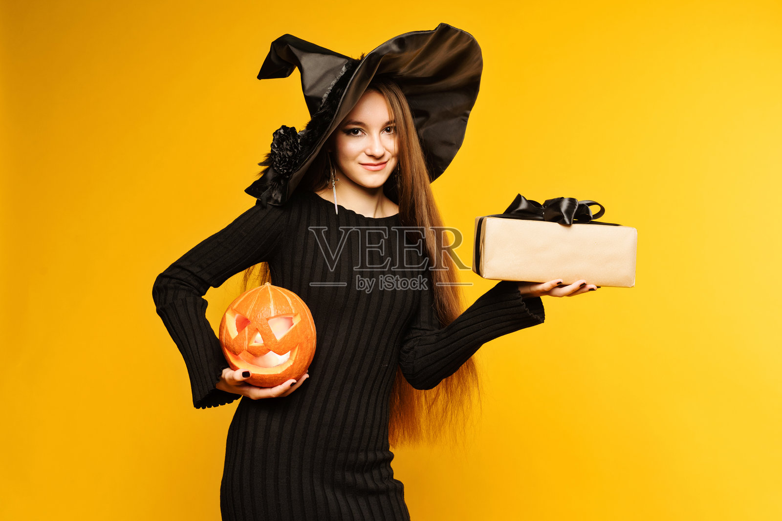 年轻女性穿着黑色连衣裙和帽子,手持南瓜和礼物,背景为黄色。万圣节概念。女巫装扮的女孩。照片摄影图片