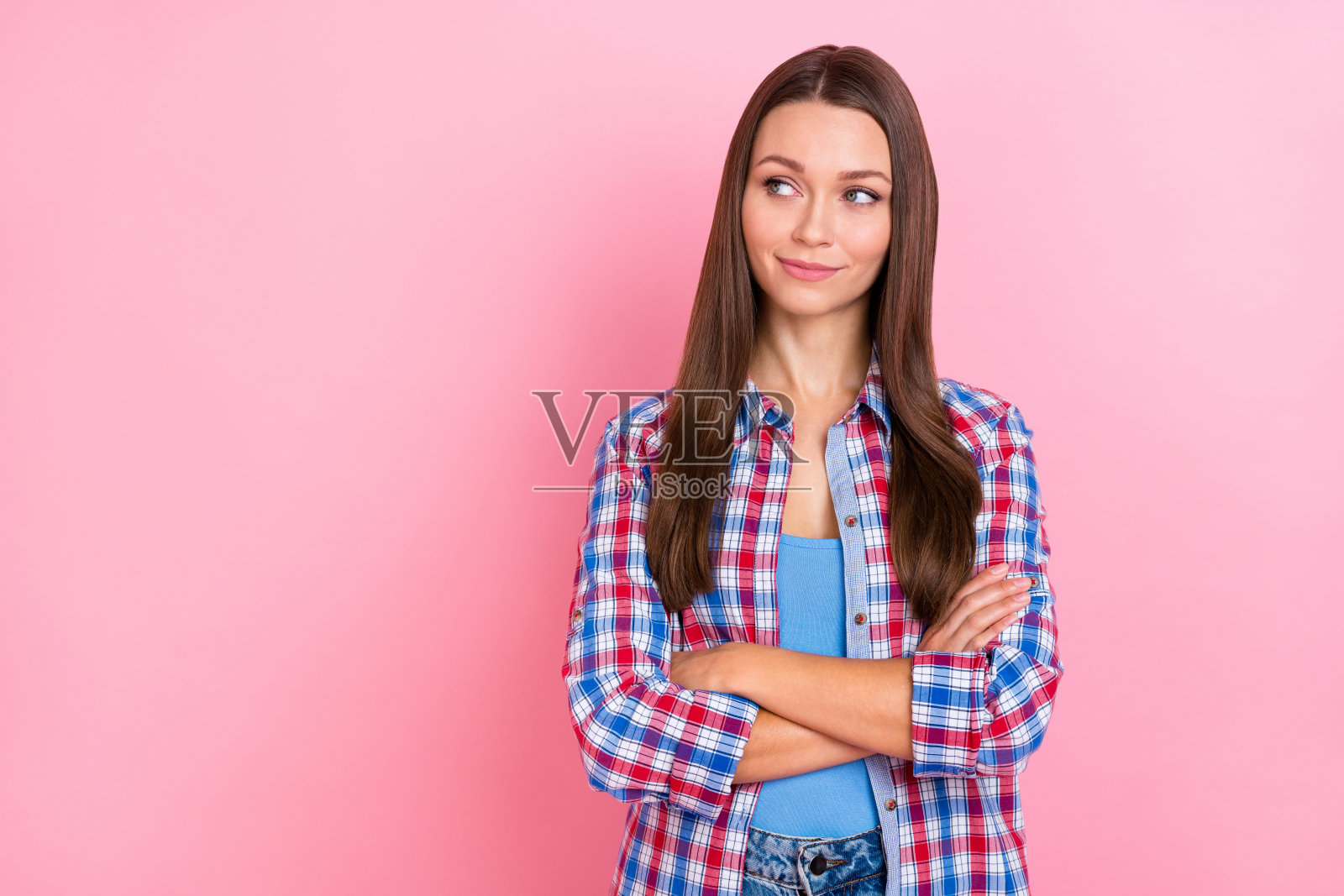 一位狡猾的棕发千禧女性双臂交叉,凝视空旷的空间,穿着蓝红相间的衬衫,背景为柔和的粉色。照片摄影图片