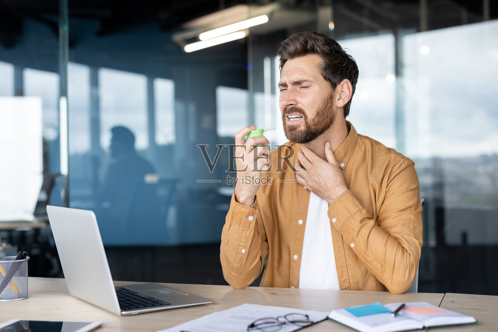 一名男子在办公室桌前使用喉咙喷雾治疗感冒或喉咙痛。照片摄影图片