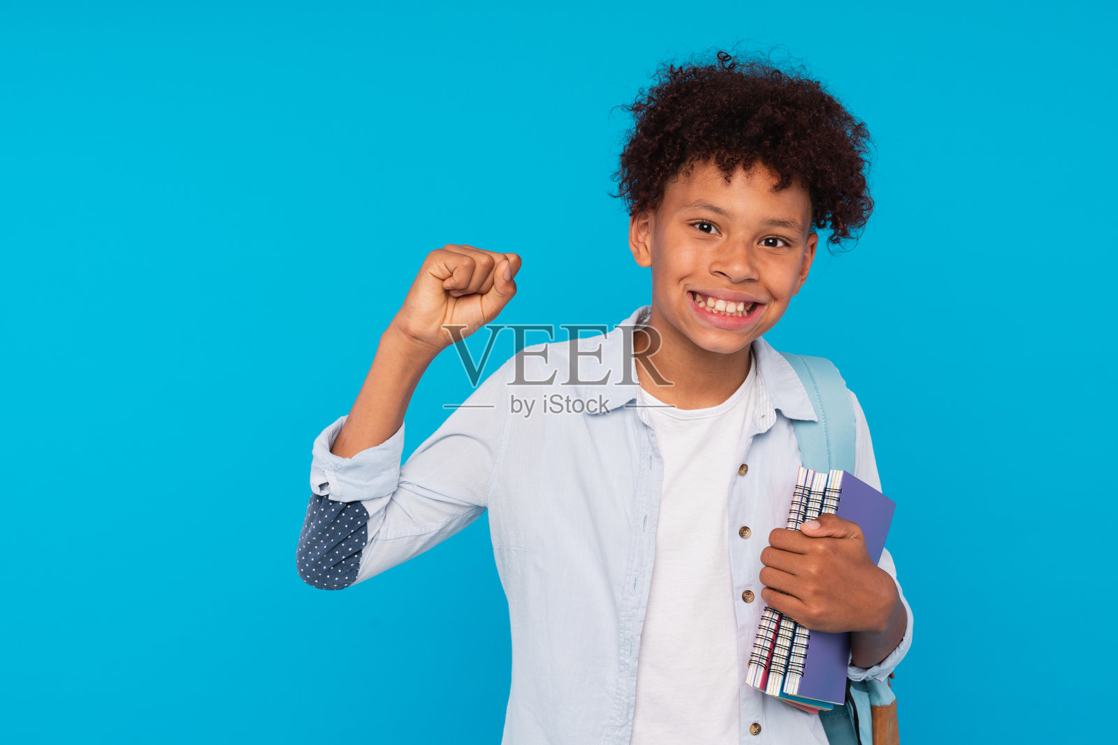 微笑的非裔美国小学生男孩,背着书包,手里拿着书本和笔记本,做出胜利手势庆祝成功,独自站在蓝色背景前,留有空白空间。照片摄影图片