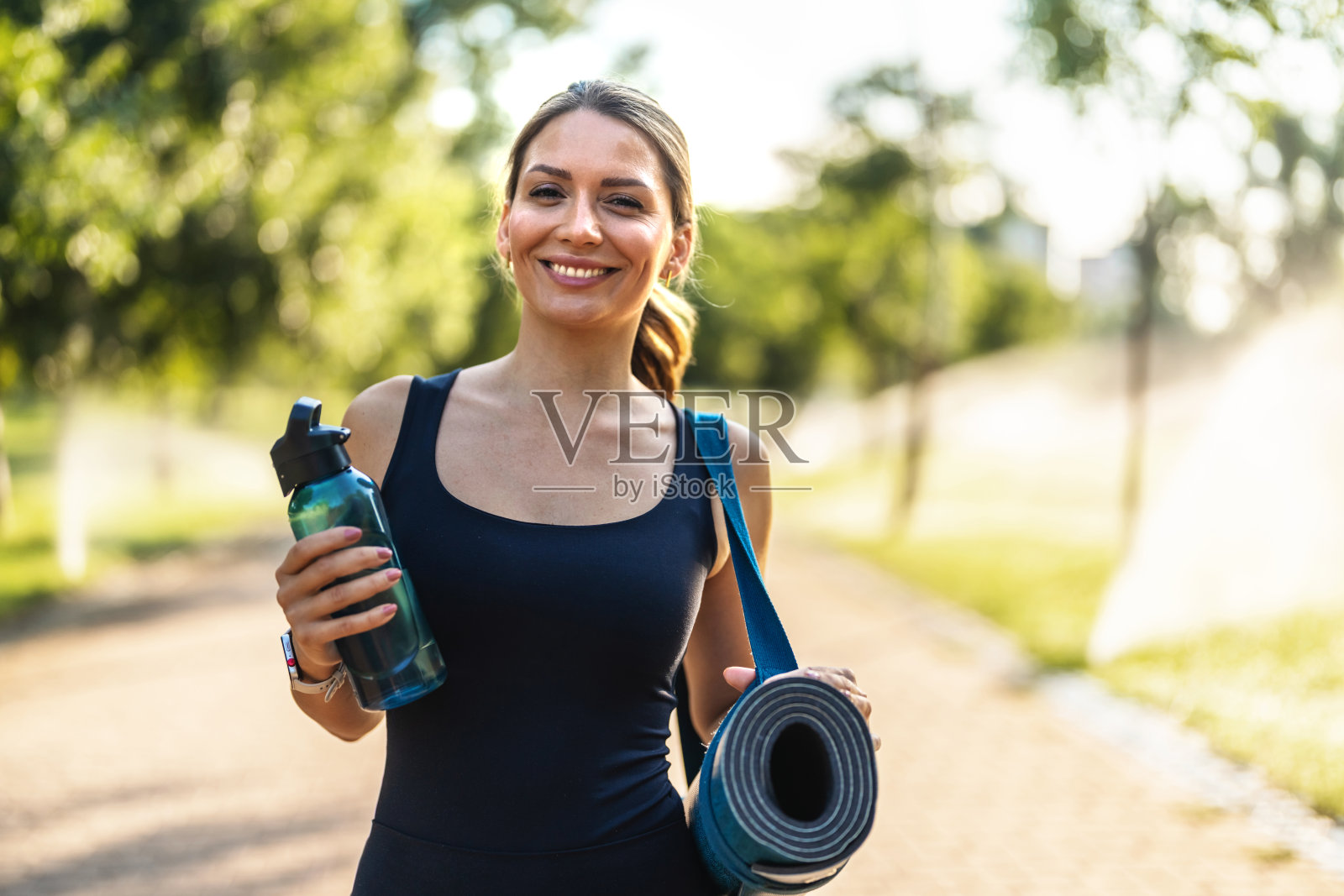 在公园里走路的女性,手里拿着瑜伽垫,准备进行健康锻炼,思考或准备进行普拉提。下面,瑜伽爱好者在大自然中思考,带着器材进行健康日常,开始锻炼或享受新鲜空气。照片摄影图片