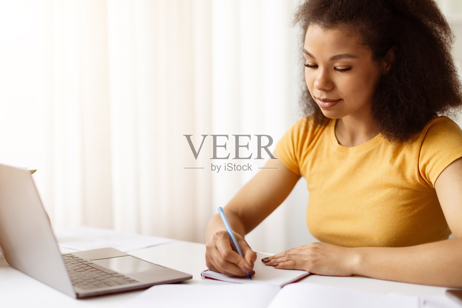 专注的非裔美国女性在笔记本上写笔记,同时在笔记本电脑前学习照片摄影图片