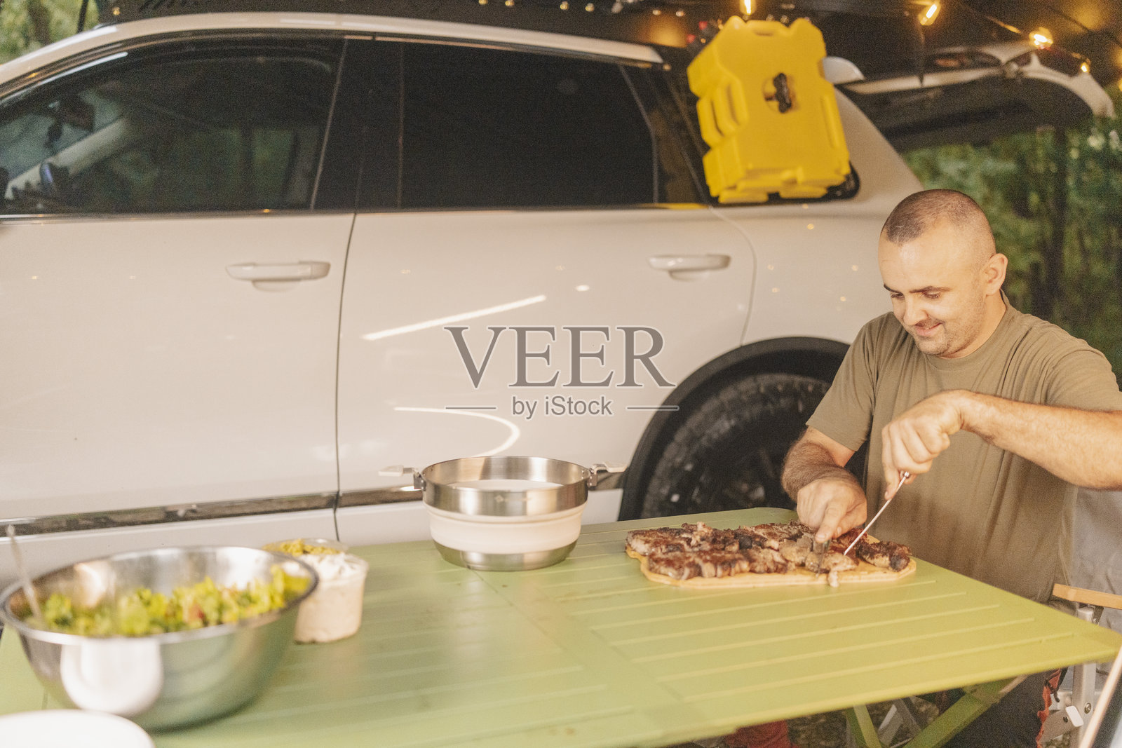 男人在汽车旁的露营地切割烤肉,周围是傍晚的灯光照片摄影图片