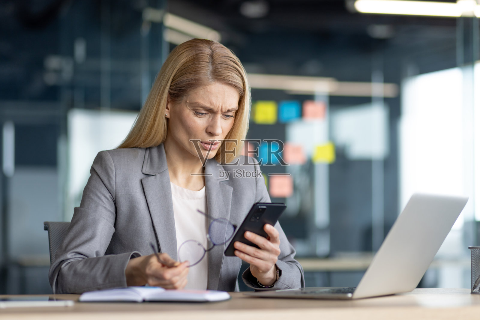 困惑的女商人一边工作一边看着智能手机,身处现代办公空间照片摄影图片