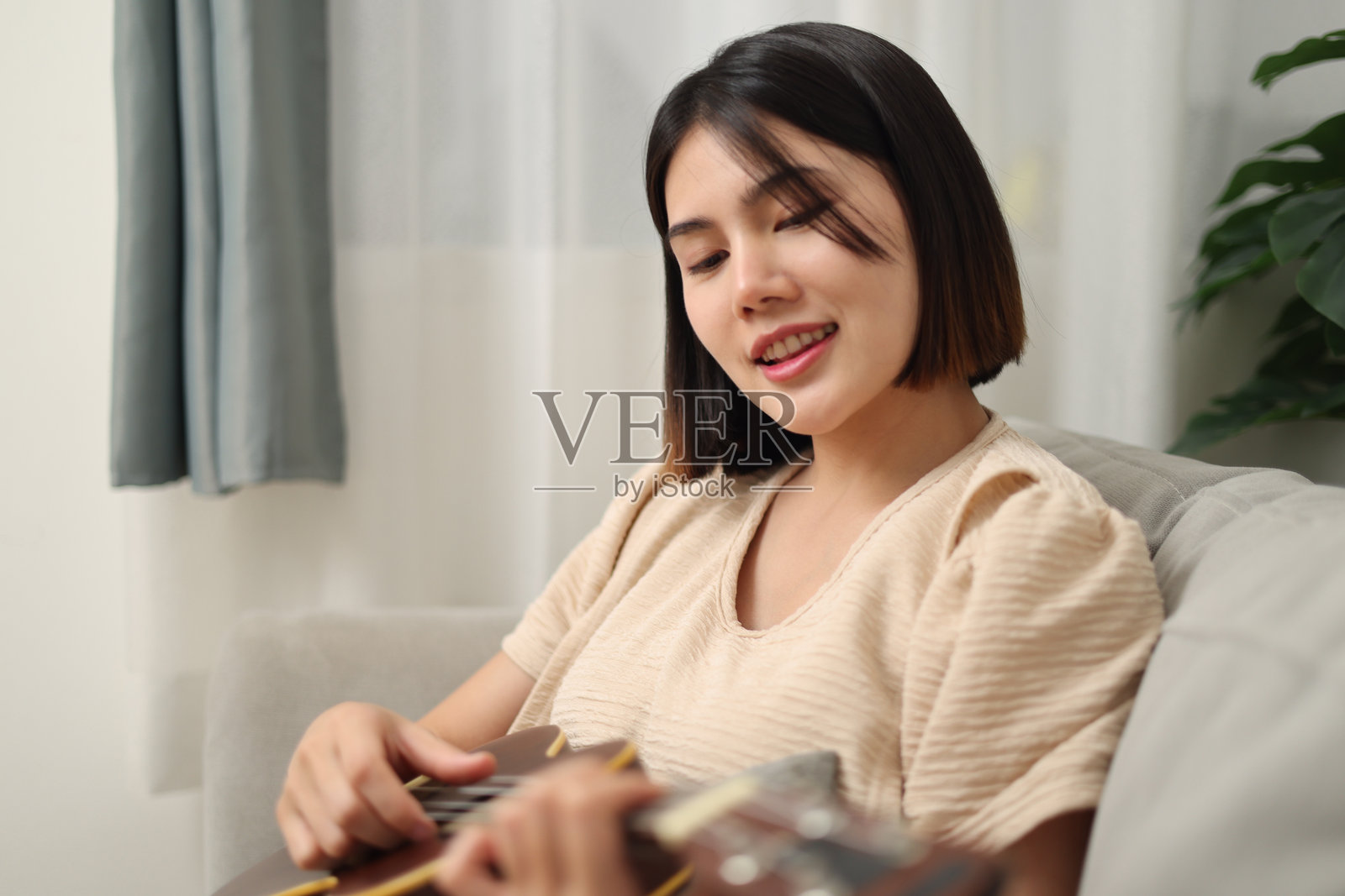 年轻的亚洲女性在家里的沙发上享受弹尤克里里。照片摄影图片