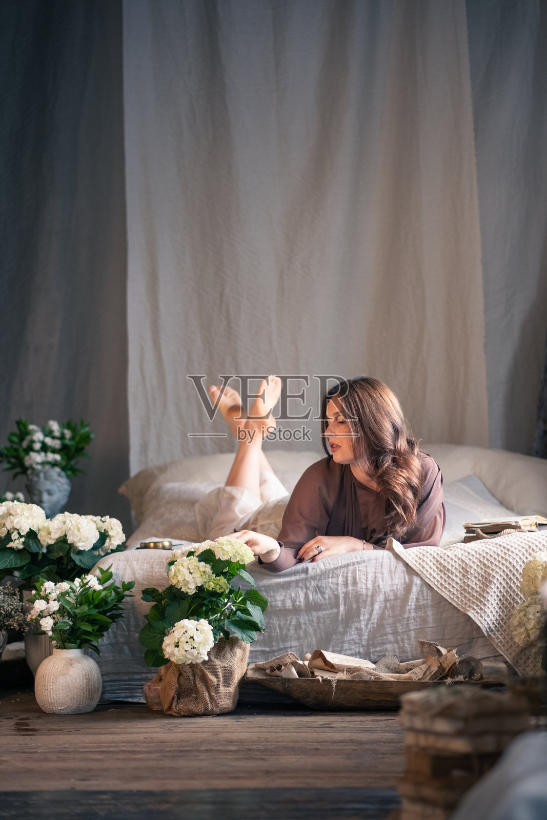 美丽的年轻女性在复古风格的房间里床上摆姿势,周围有很多花。背景是工业风格的砖墙。照片摄影图片