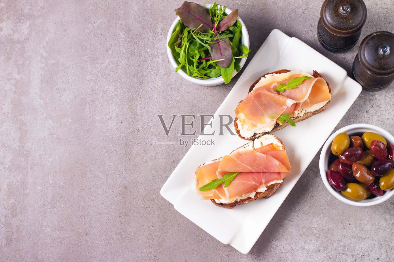 两片意式开胃面包,上面放有火腿、瑞可塔奶酪和芝麻菜,搭配橄榄和香料装饰。照片摄影图片