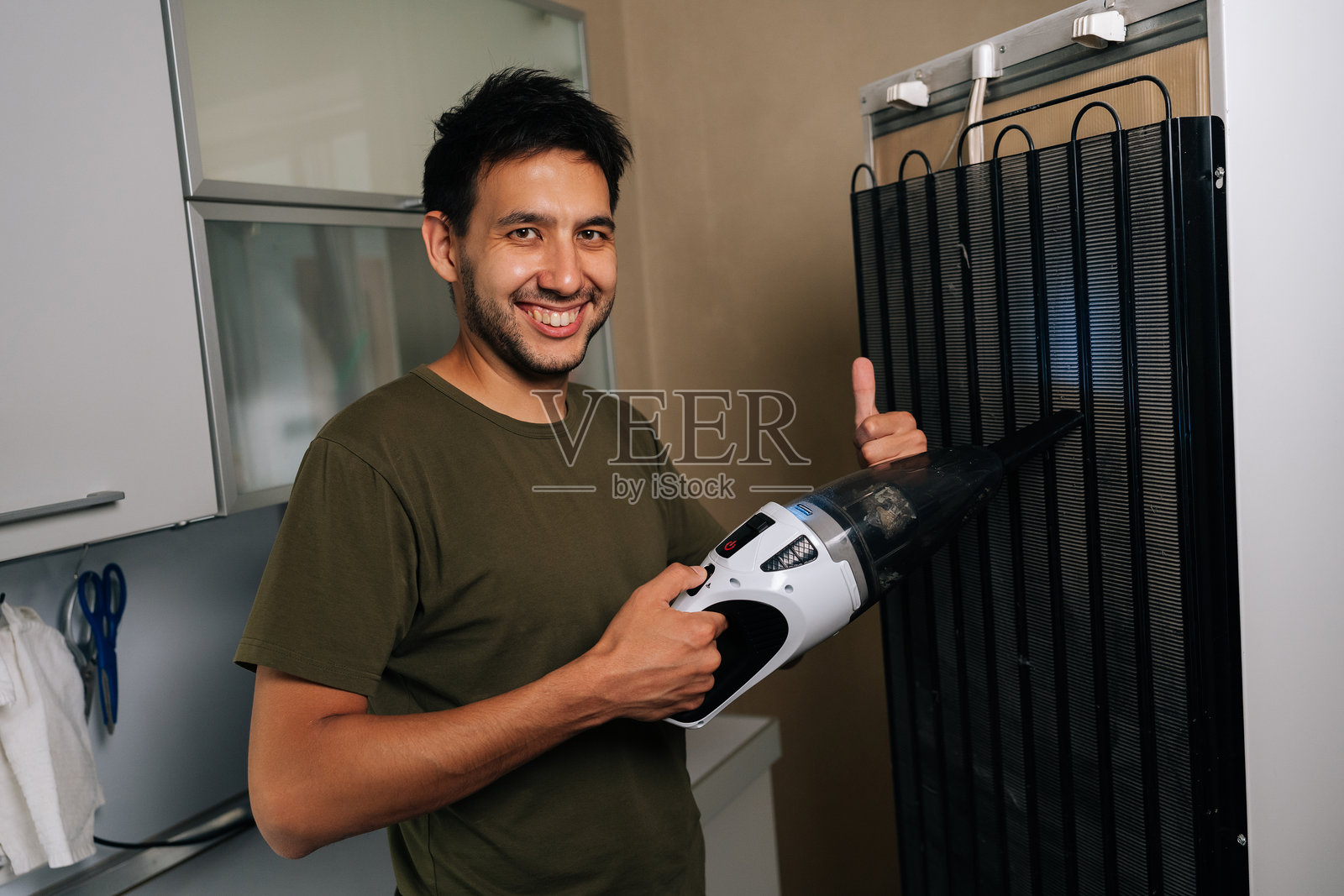 友好的清洁服务员工正在用便携式吸尘器清洁冰箱的冷却线圈,并做出竖起大拇指的手势。照片摄影图片