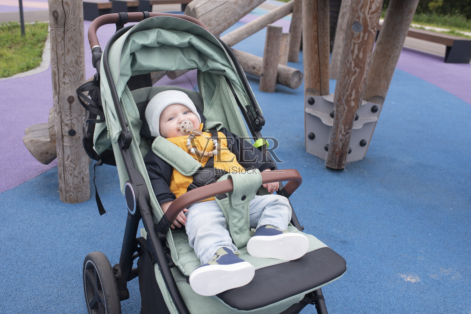 小男孩穿着夹克和帽子坐在游乐场的绿色婴儿车里照片摄影图片