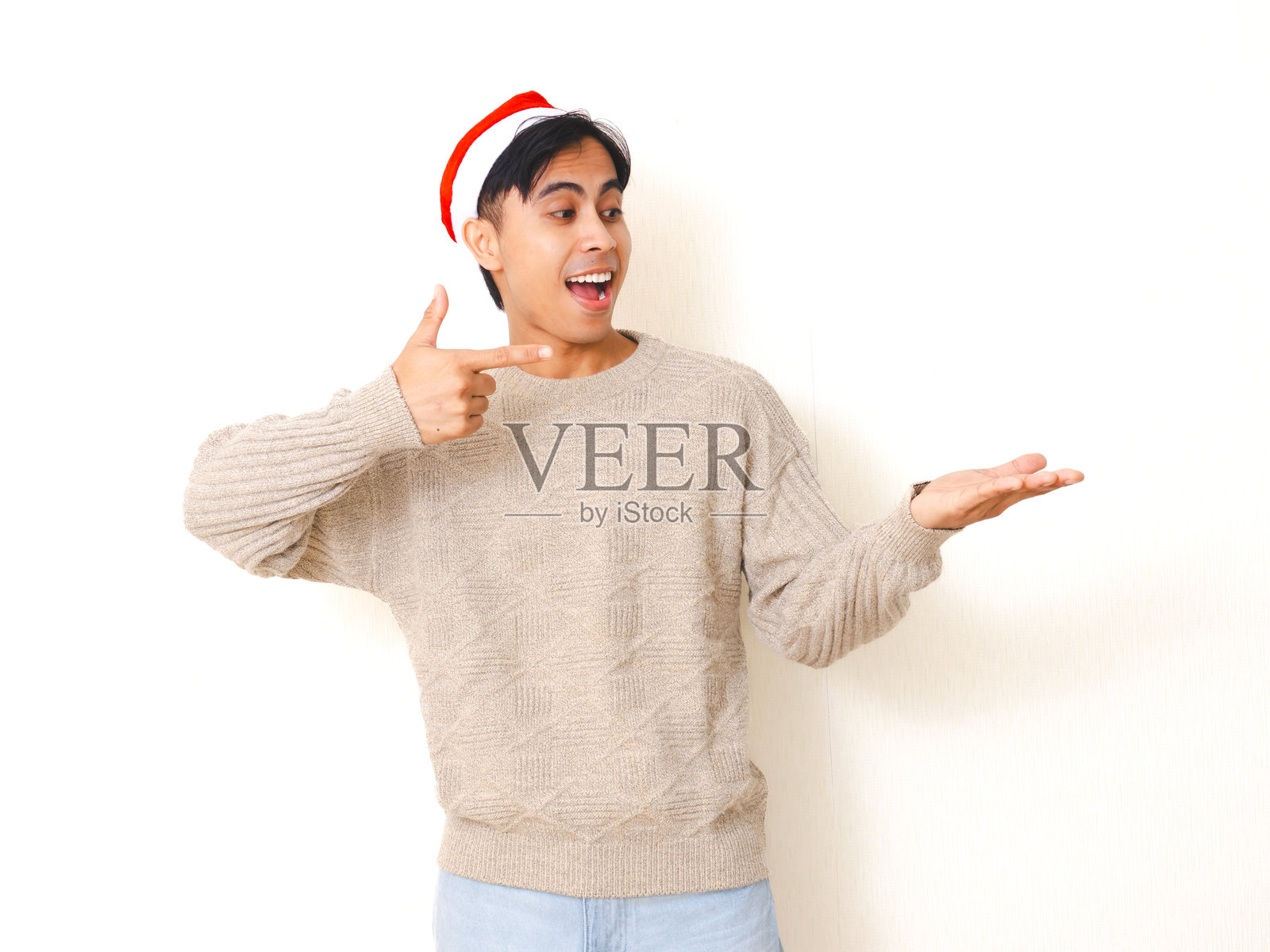 戴着圣诞帽的年轻男子兴奋地指向空空的掌心,背景干净的白色,非常适合节日和庆典概念。照片摄影图片