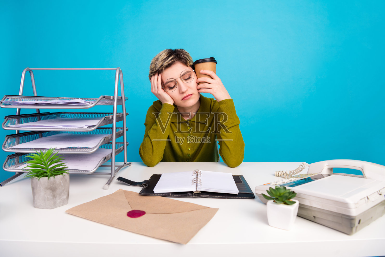 疲惫的女商人在工作区手持咖啡,背景是文件、植物和复古电话,蓝色背景照片摄影图片