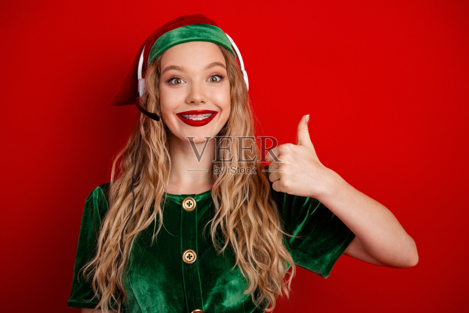 一位身着节日精灵服装的快乐年轻女子,在充满活力的季节性红色背景下竖起大拇指。照片摄影图片