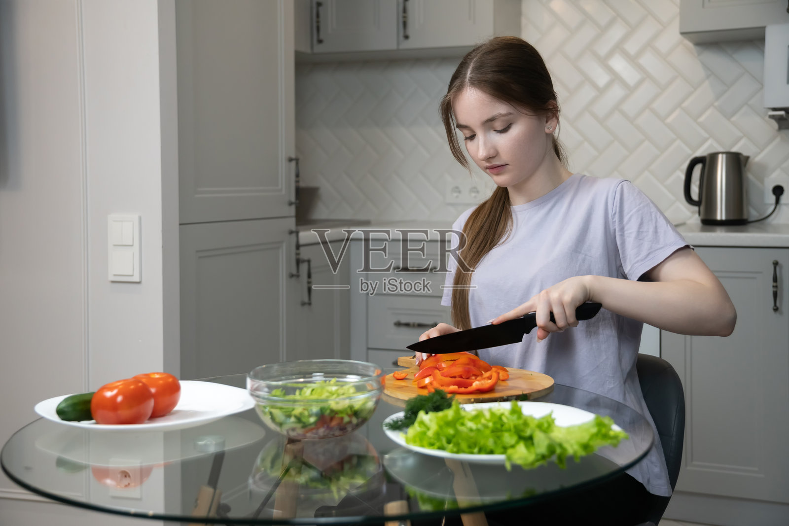 年轻女子坐在玻璃厨房桌旁,在木质砧板上仔细切红甜椒,周围摆放着新鲜的生菜、莳萝和其他沙拉配料,准备制作健康餐食。照片摄影图片