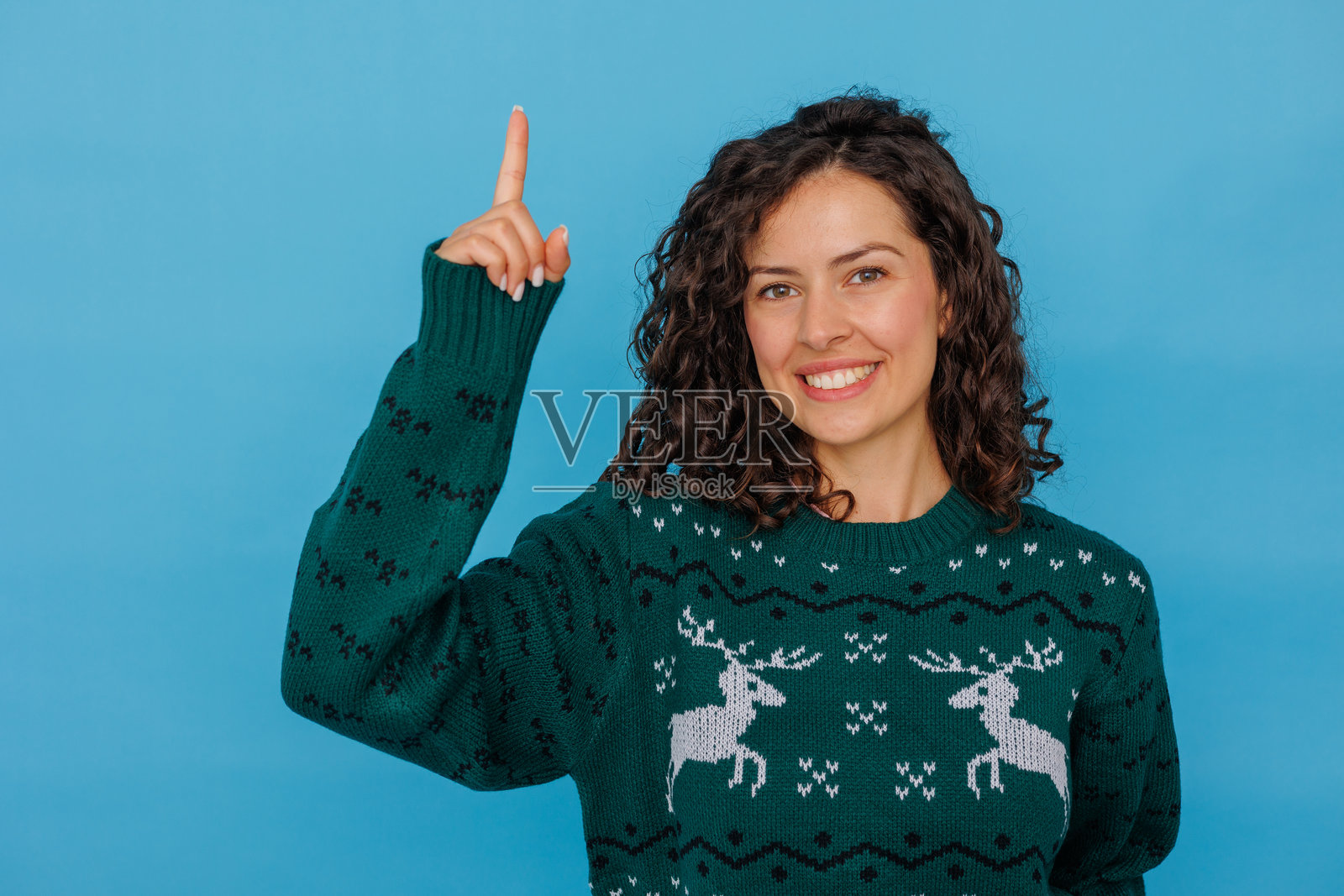 一位微笑着的女士穿着圣诞毛衣,手指向上,在蓝色背景上展示一个想法或方向。照片摄影图片