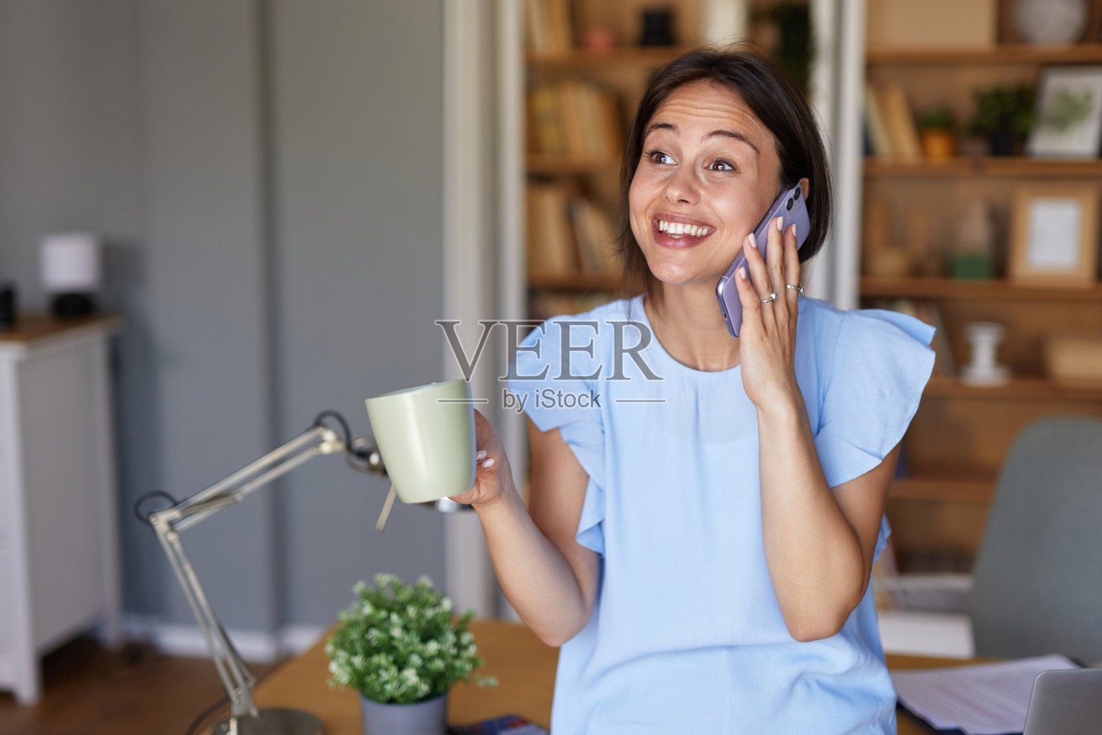 快乐的年轻女子在家办公,一边喝咖啡一边打电话。照片摄影图片