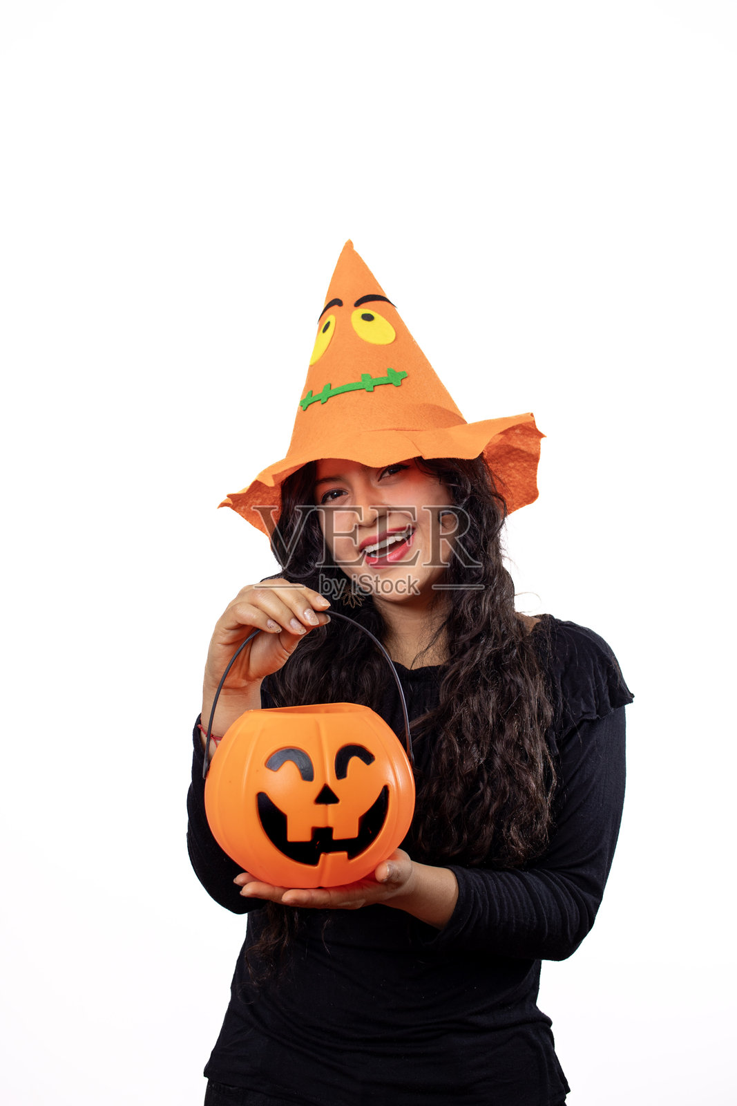 一位年轻的西班牙裔女性,戴着节日哈利波特帽,微笑着拿着一个南瓜形状的糖果桶,在白色背景的影棚肖像中快乐地摆姿势。照片摄影图片