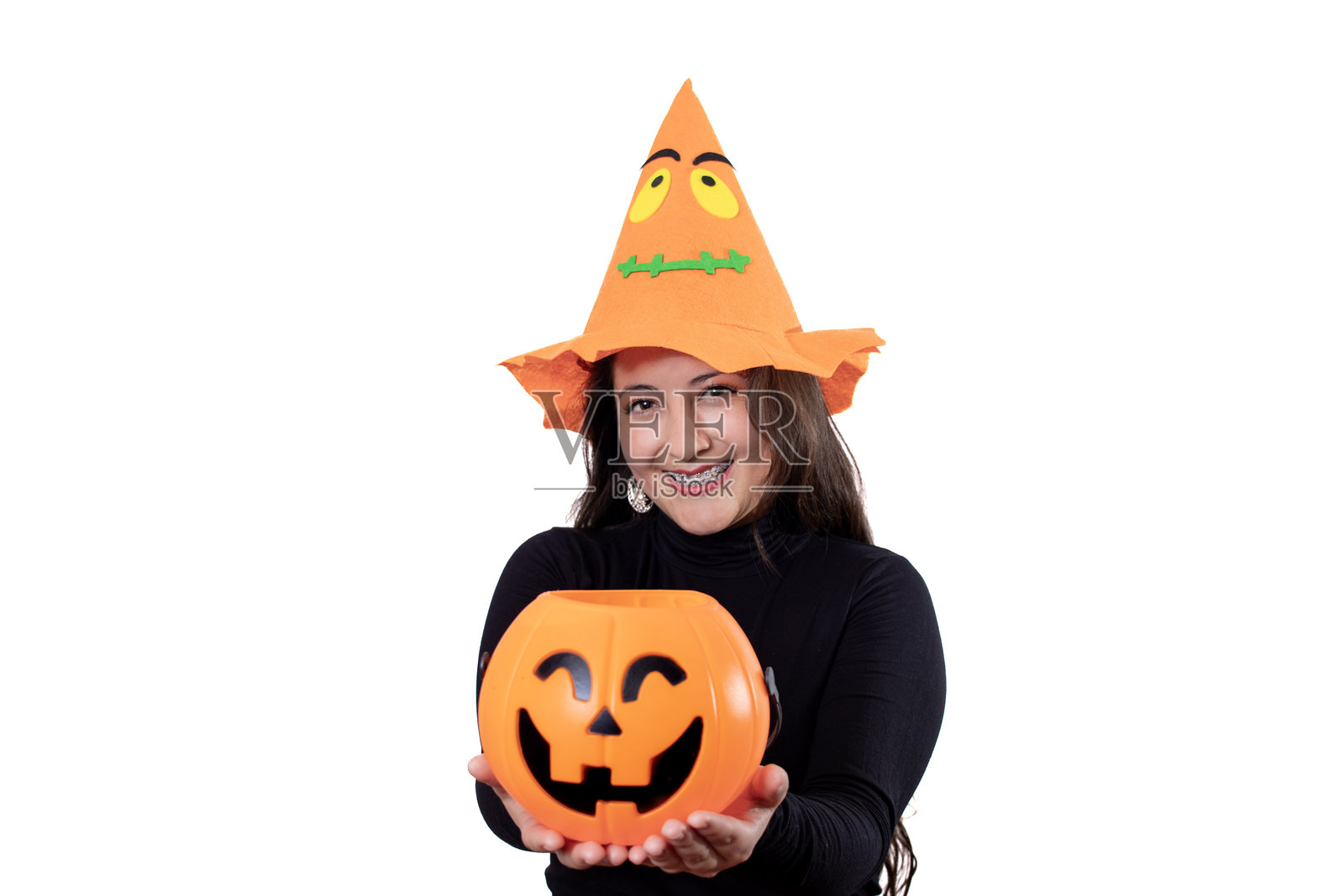 年轻女子穿着万圣节服装,戴着橙色南瓜帽,穿着黑色衬衫,手持塑料南瓜灯糖果盒,看向镜头照片摄影图片