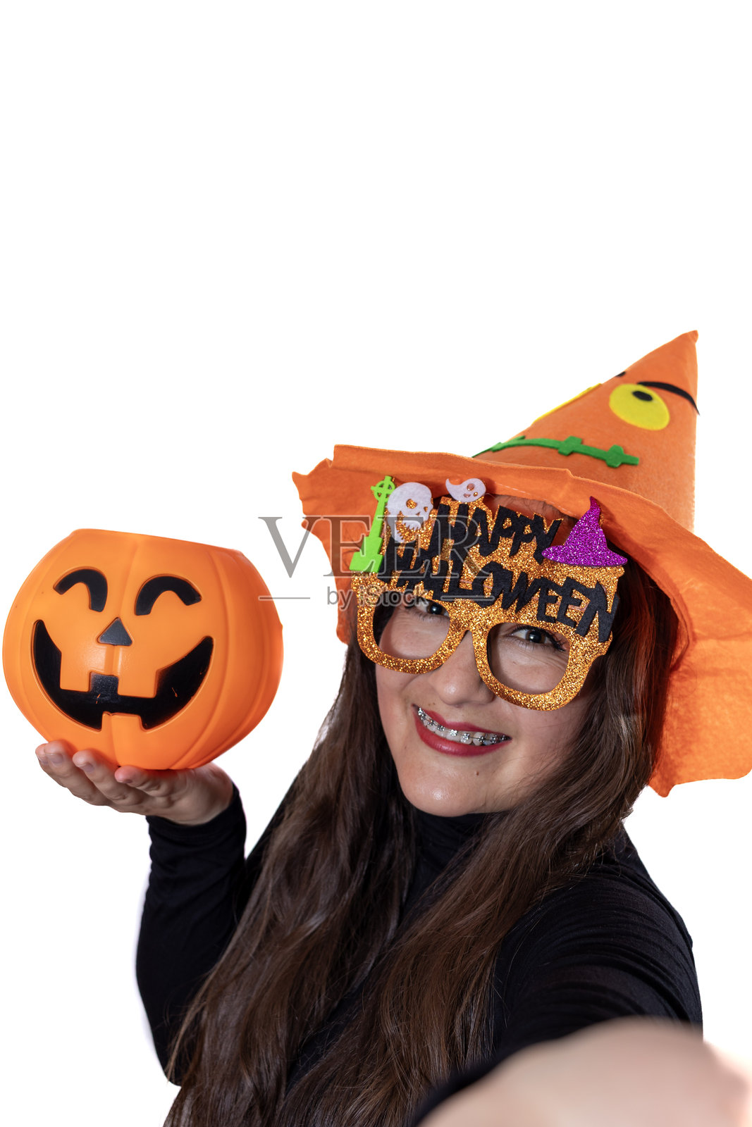 年轻女子享受万圣节派对,戴着巫师帽,戴着节日快乐的万圣节眼镜,手里拿着南瓜灯糖果桶,自信地笑着自拍照片摄影图片
