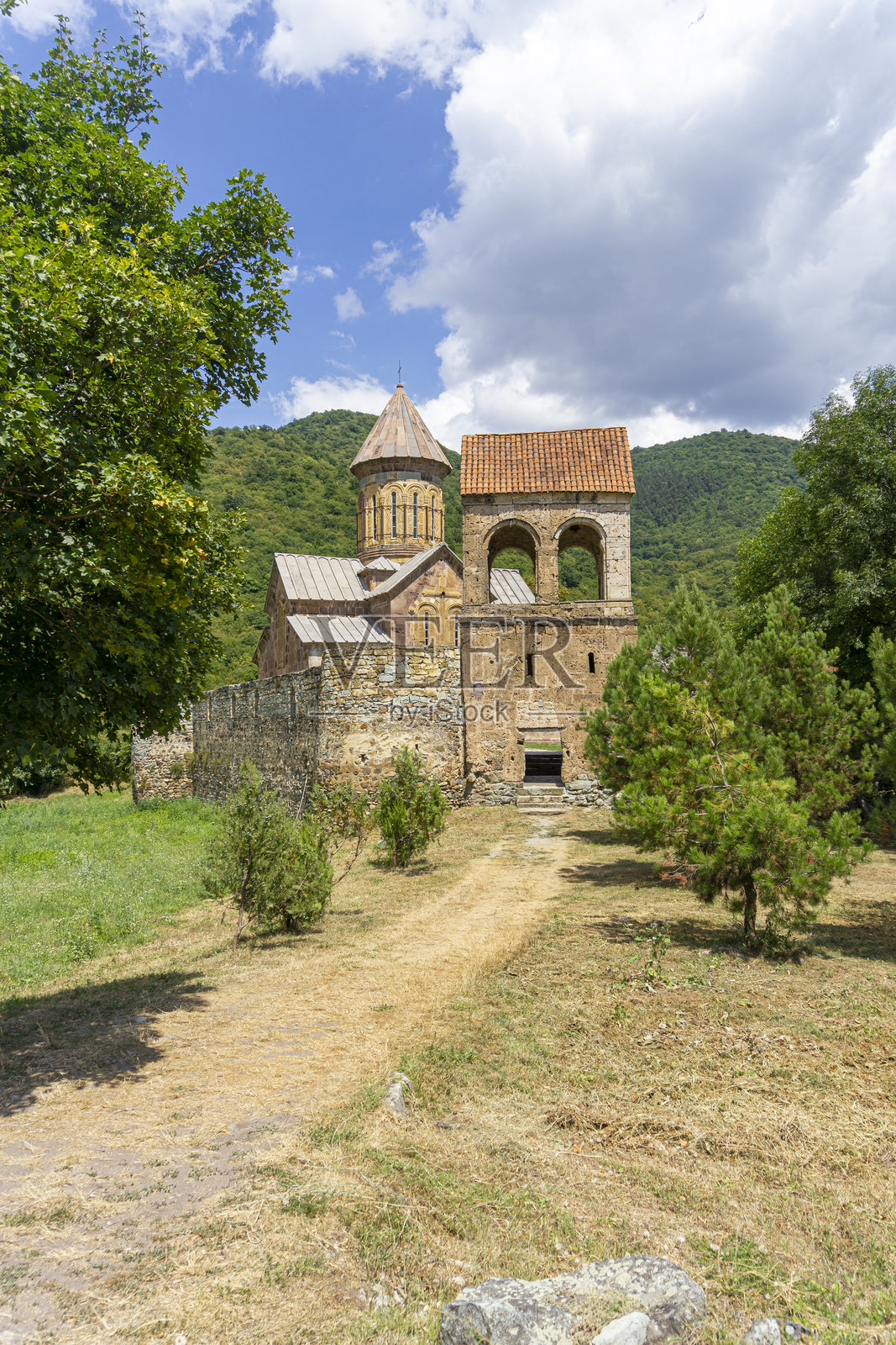 一条长满褪色野草的土路,穿过高耸的钟楼,通向寺院庭院的入口,可以看到教堂的圆顶。皮特雷蒂修道院。格鲁吉亚。照片摄影图片
