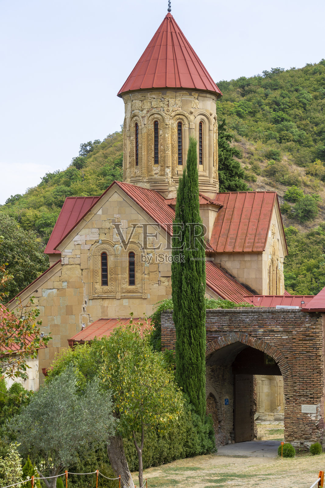 一座带有圆顶和棕色屋顶的石头教堂坐落于峡谷中,砖墙上有一个拱形通道。背景是覆盖着灌木和树木的山丘,以及蓝色的天空。贝塔尼亚修道院。照片摄影图片