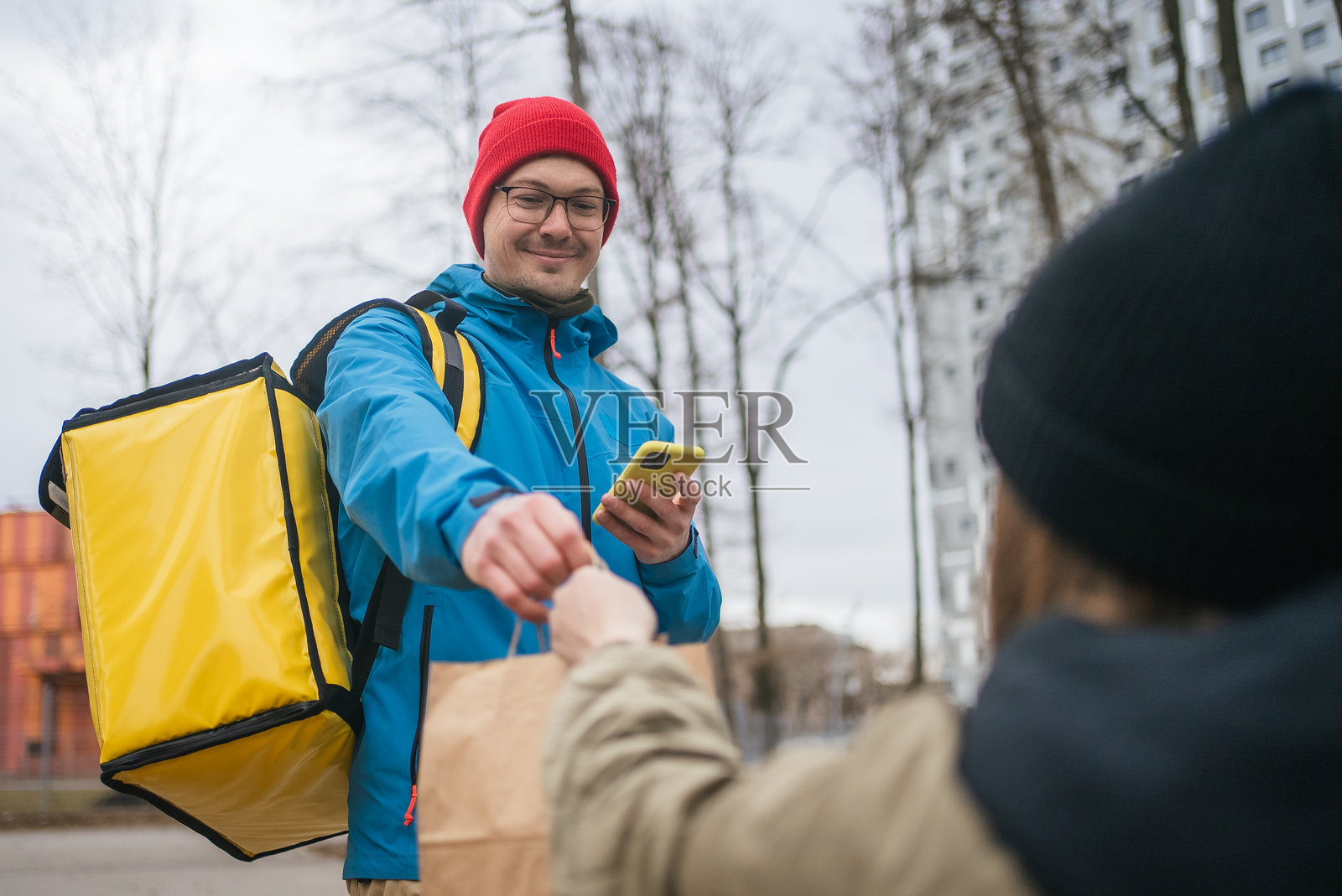 送餐员在寒冷天气中,背着保温背包,手持智能手机和纸袋,穿梭于城市街道,递送外卖餐食。照片摄影图片