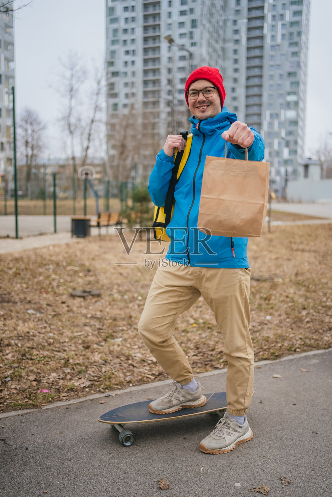 穿着红色帽子和蓝色夹克的配送员,脚踩长板,手提装着外卖食品的纸袋,为顾客配送。照片摄影图片