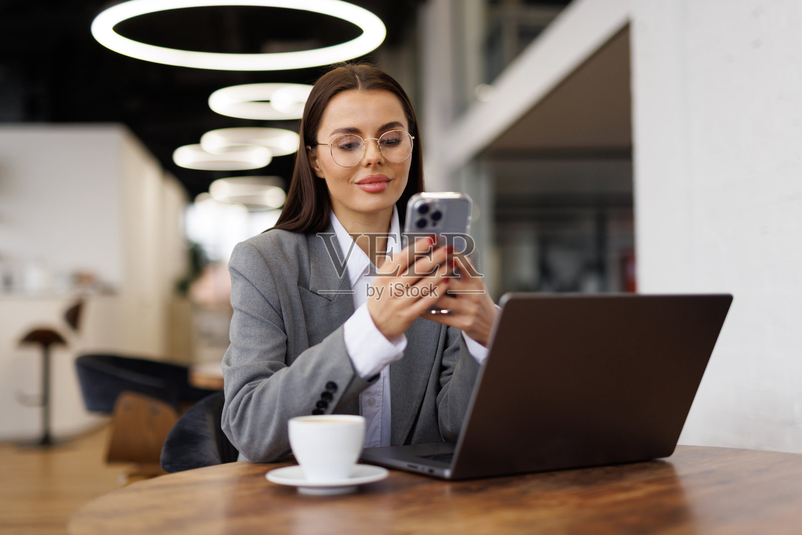 年轻女子坐在咖啡馆的木桌旁,一边喝咖啡一边使用智能手机。照片摄影图片