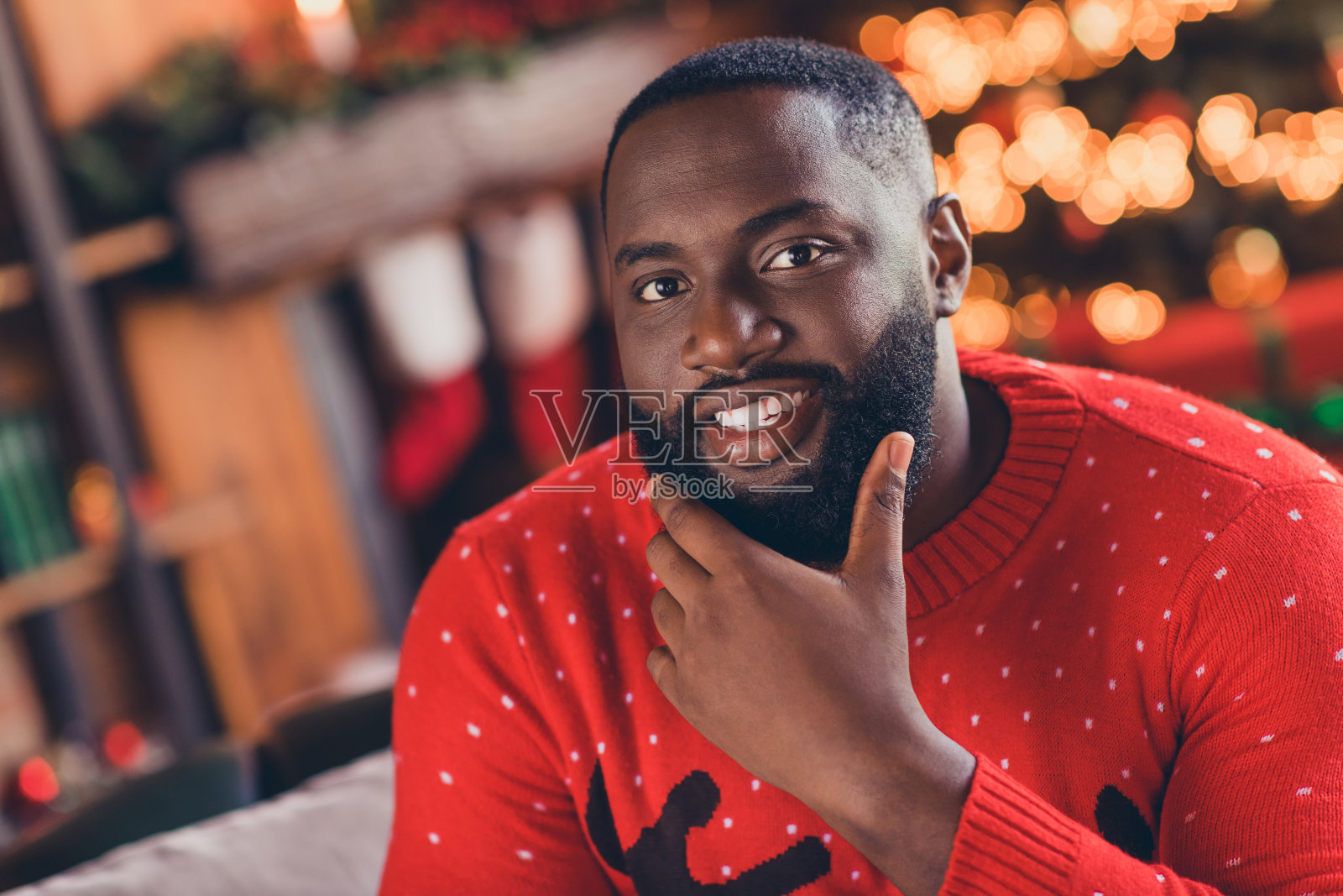 梦幻可爱深肤色男性,身穿印花套头衫,面带笑容,手臂搭在下巴上,在室内房屋房间里。照片摄影图片