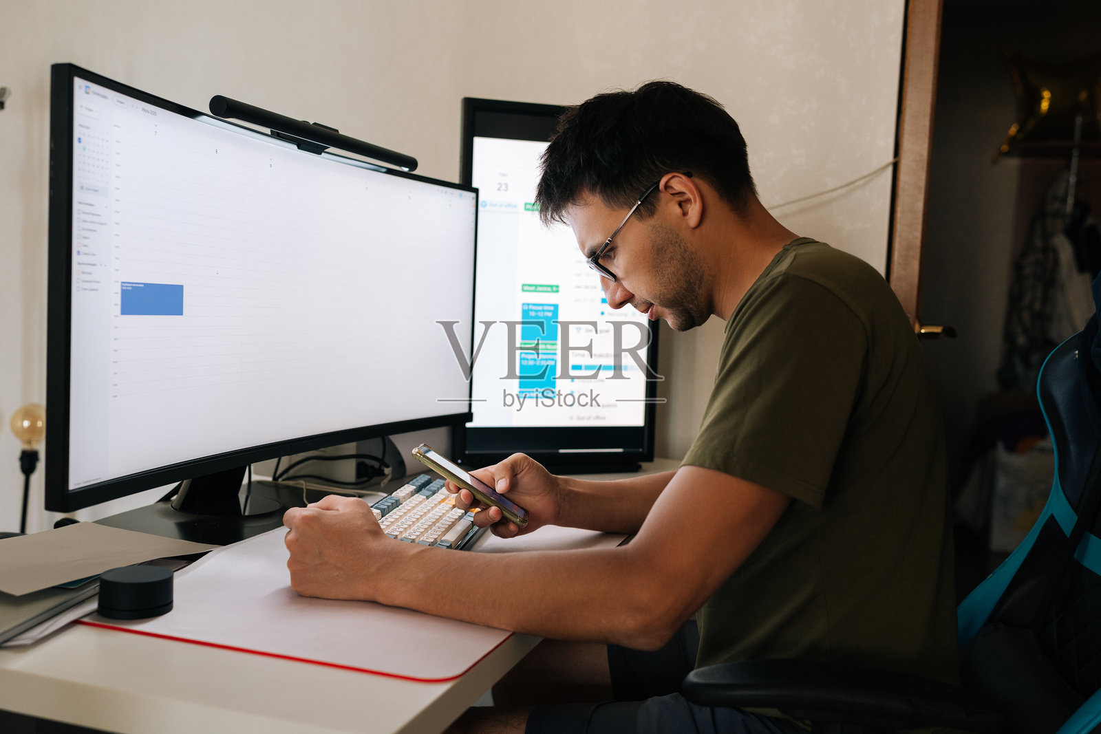 程序员在家办公,坐在书桌前,两台电脑屏幕前,用手机查看日历并安排会议。照片摄影图片