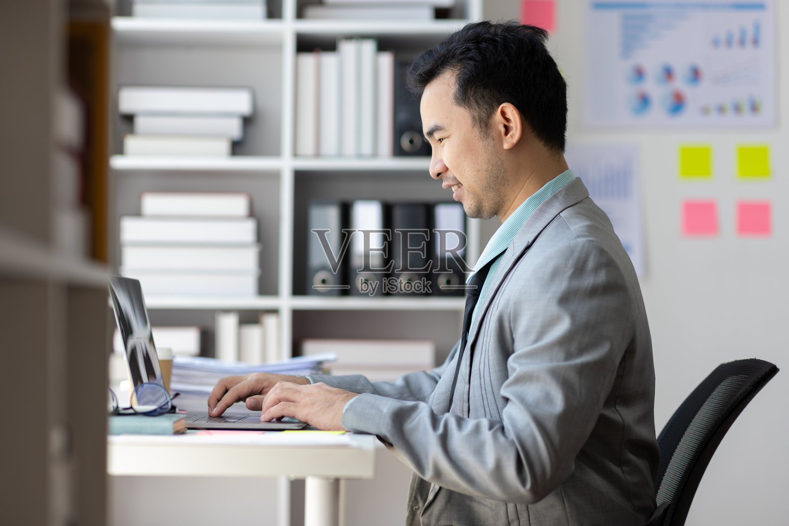 一位亚洲商人正在现代办公室里对着笔记本电脑微笑工作,旁边有书架,墙上有便利贴。照片摄影图片