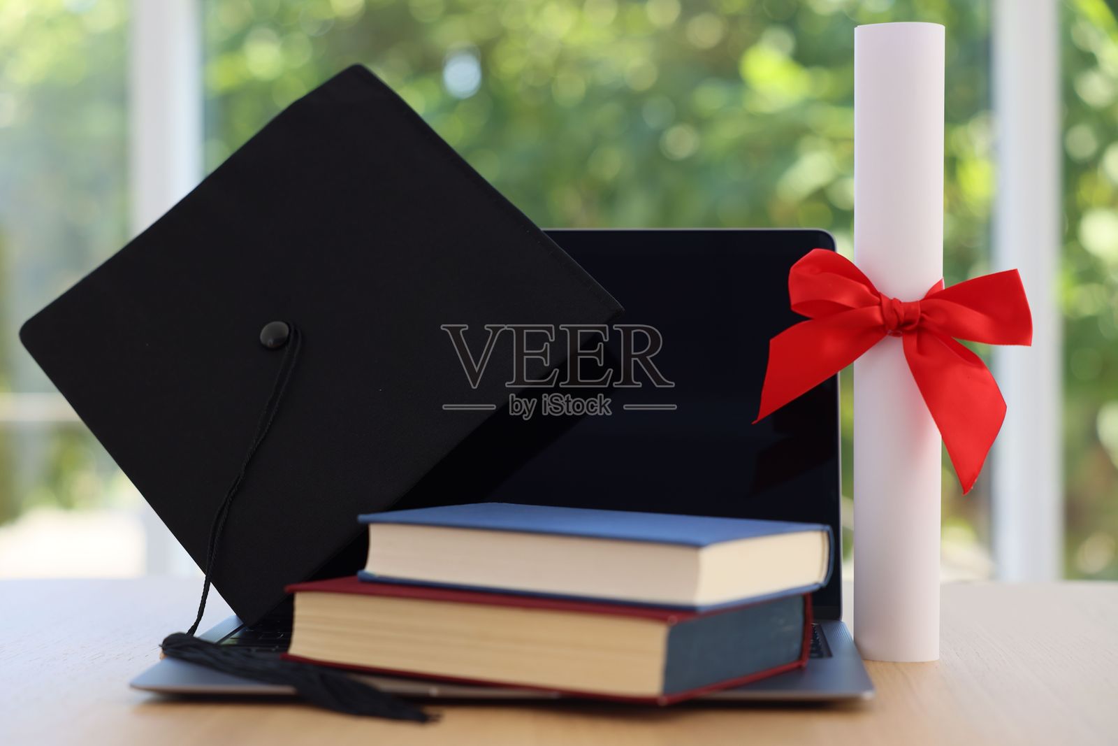 毕业帽、笔记本电脑、书籍和文凭放在木桌上,背景是模糊的绿色,特写。照片摄影图片