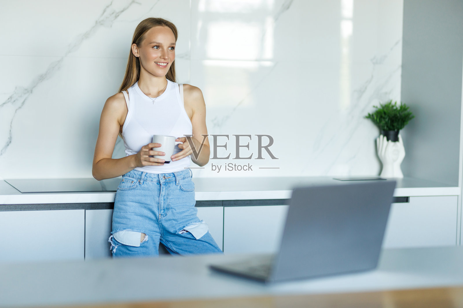 一位美丽的年轻女子在家中厨房的台面上,一边喝咖啡一边用笔记本电脑工作,脸上带着笑容。照片摄影图片
