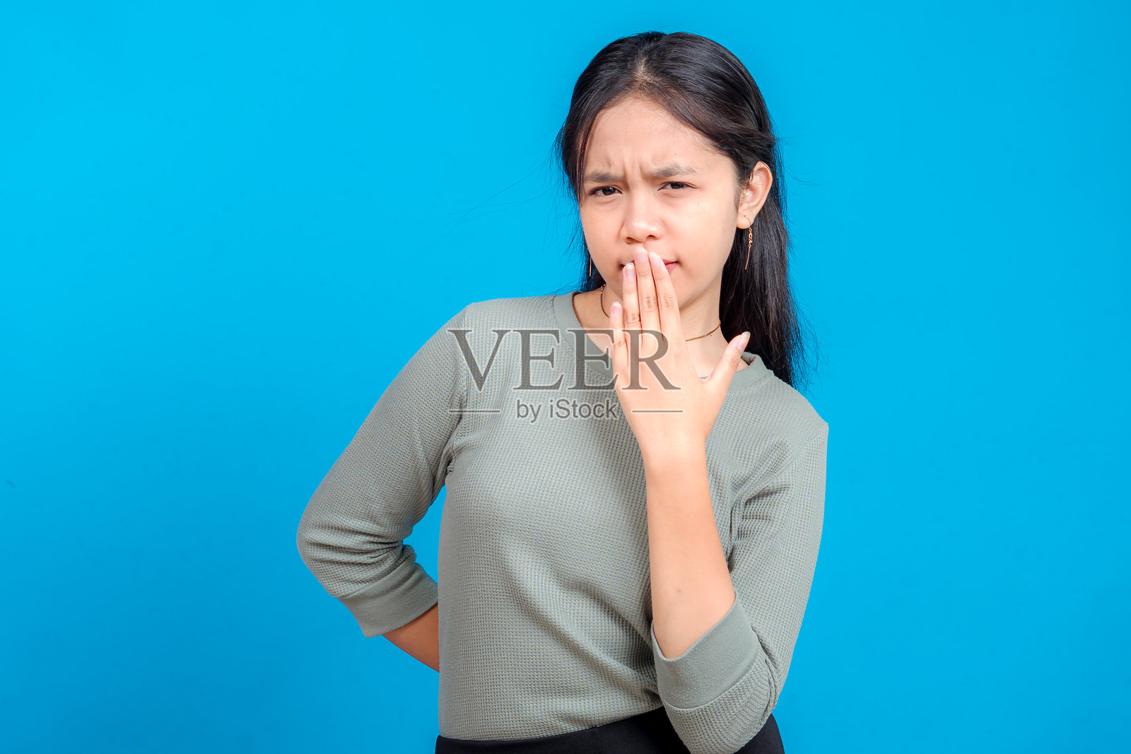身处蓝色背景前,一位年轻亚洲女性做出怀疑和不信任的表情,挑起眉毛,用手捂住嘴巴,工作室拍摄。照片摄影图片