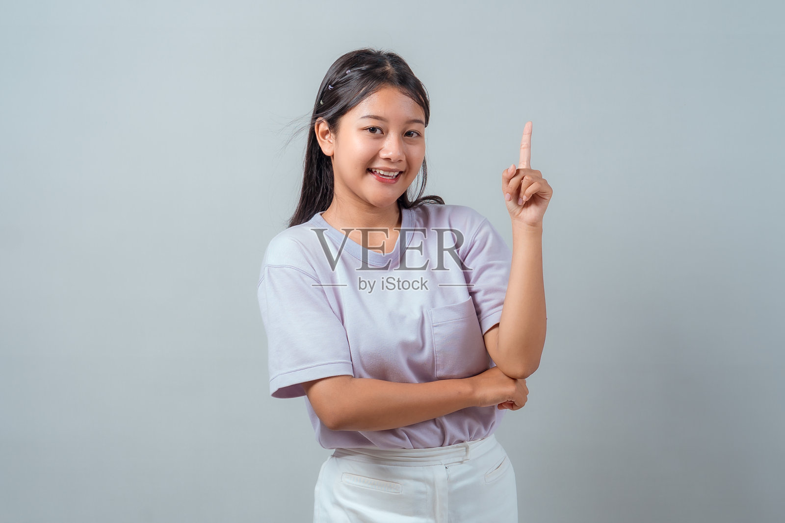一位年轻的亚洲女孩,面带微笑,身穿浅紫色T恤和白色裤子,自信地用一根手指向上指着,背景纯净,有复制空间。照片摄影图片