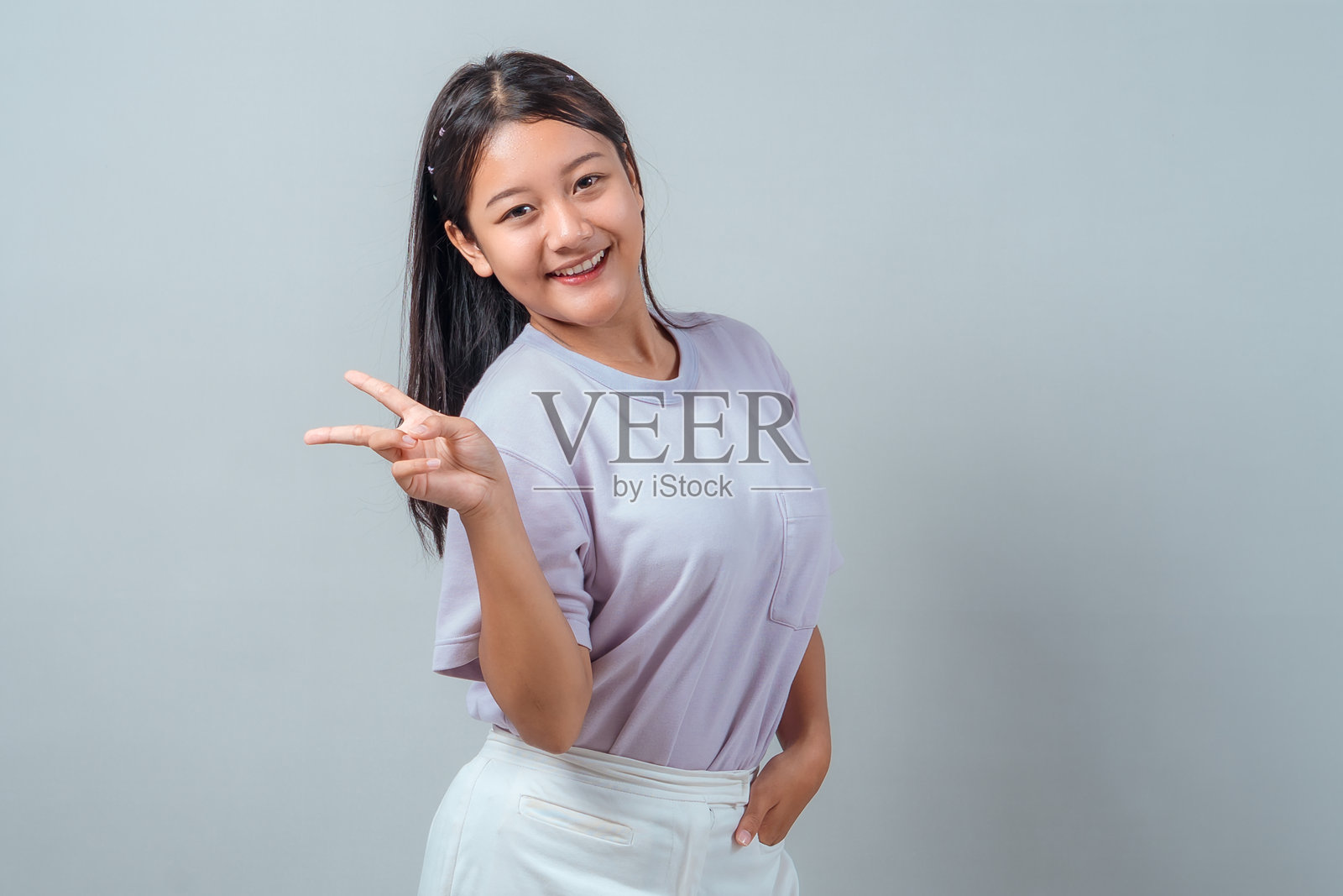 一位年轻的亚洲女性微笑着竖起剪刀手,穿着休闲服,在工作室的灰色背景前自信地摆出俏皮的姿势。照片摄影图片
