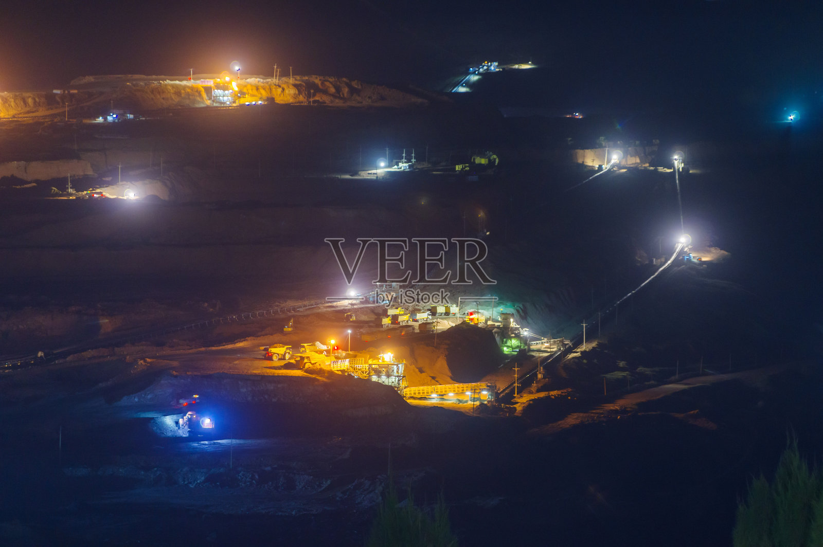 泰国兰邦府湄莫褐煤矿夜间采矿作业,重型机械和自卸卡车在广阔露天矿坑中被人工照明照亮,周围弥漫着尘土。照片摄影图片