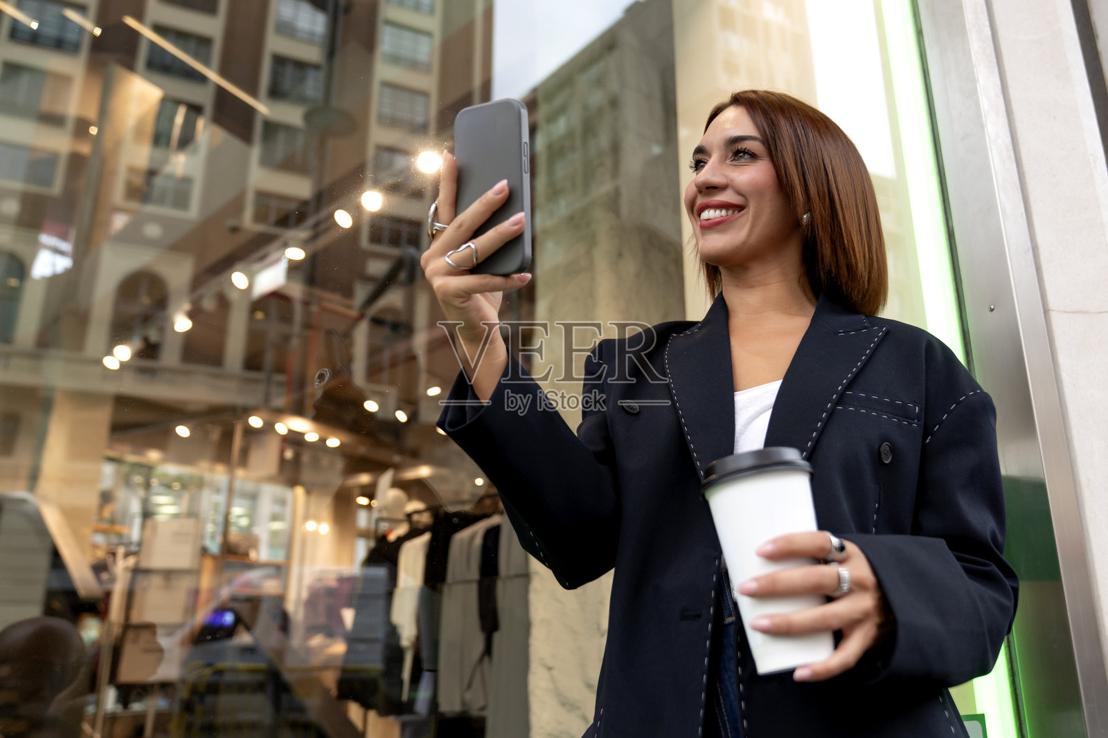 时髦的年轻女子在户外购物,一手拿着智能手机,一手拿着咖啡杯,享受着城市生活。照片摄影图片
