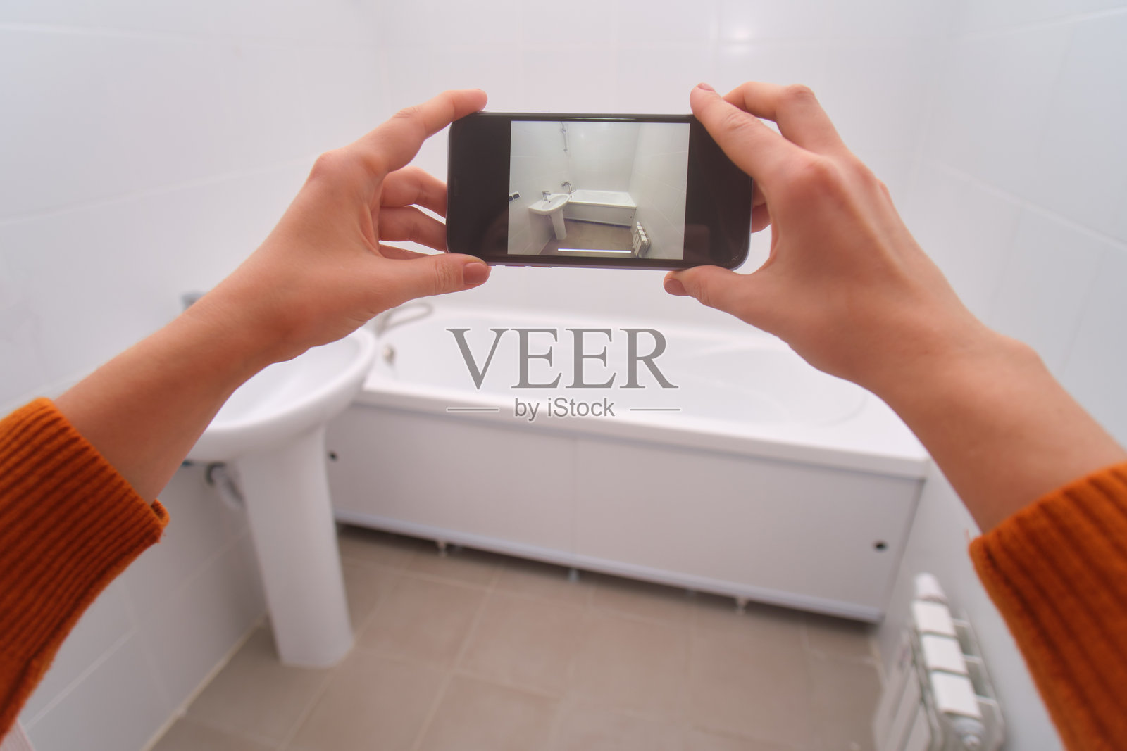 一位女士正拿着智能手机拍摄崭新白色浴室的照片,其中包括现代化的水槽和浴缸,该图片展示了翻新和室内设计概念。照片摄影图片