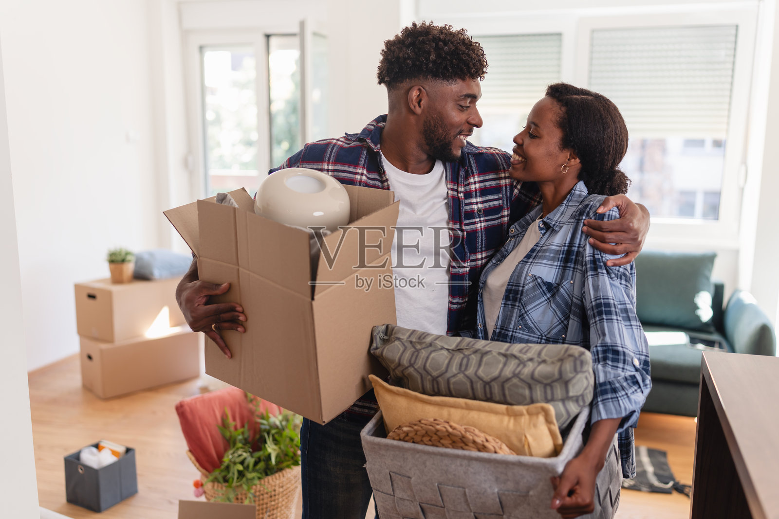 一对快乐的非洲裔美国夫妇正在拥抱,他们抱着箱子和家居装饰品,在搬家后搬进新公寓并安顿下来。照片摄影图片