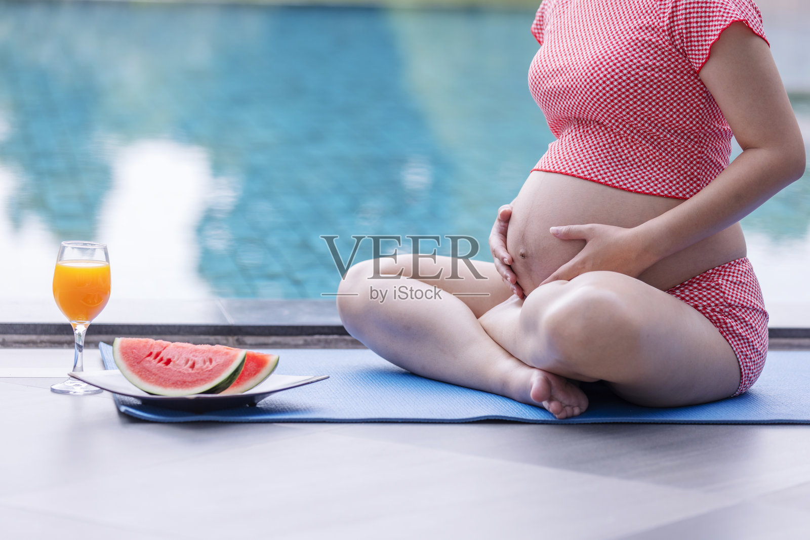 身着红色连衣裙的孕妇盘腿坐在泳池边,温柔地抚摸着自己的腹部,旁边放着西瓜和橙汁。照片摄影图片