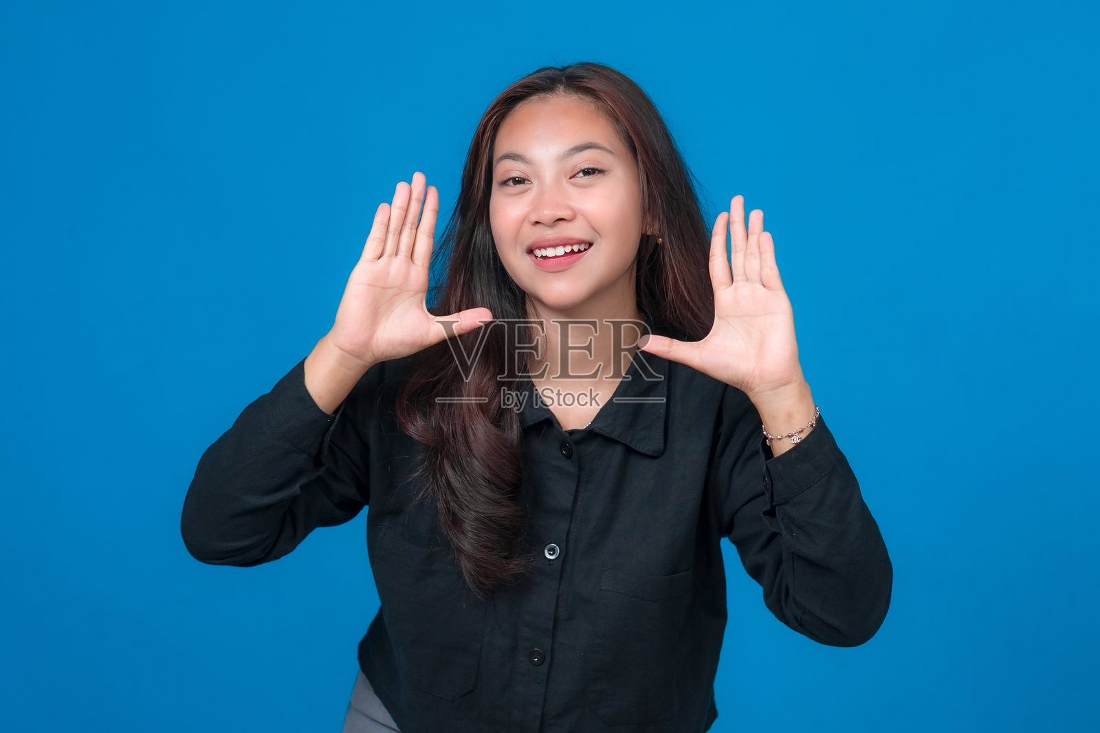 一位快乐的亚洲年轻女性,脸上带着笑容,双手举起做出有趣的姿势,身穿休闲的黑色衬衫,自信地站在明亮的蓝色背景前。照片摄影图片