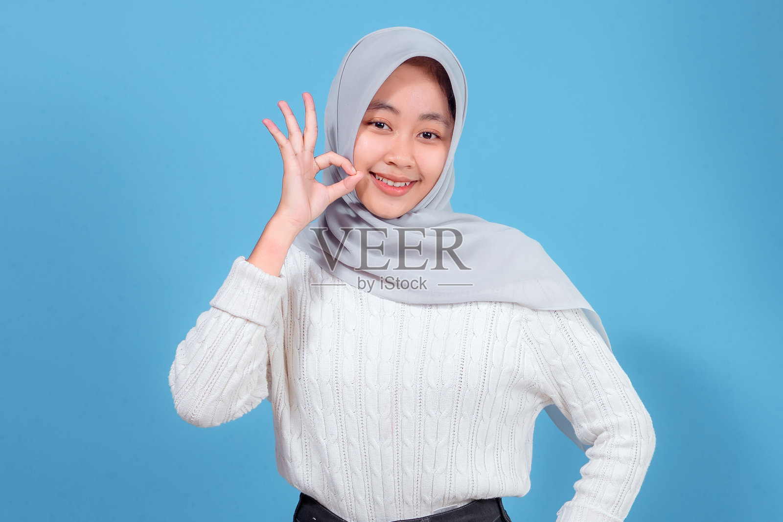 一位戴着浅灰色头巾、身穿白色针织毛衣的年轻女子,自信地竖起大拇指,摆出OK手势,背景为明亮的蓝色。照片摄影图片
