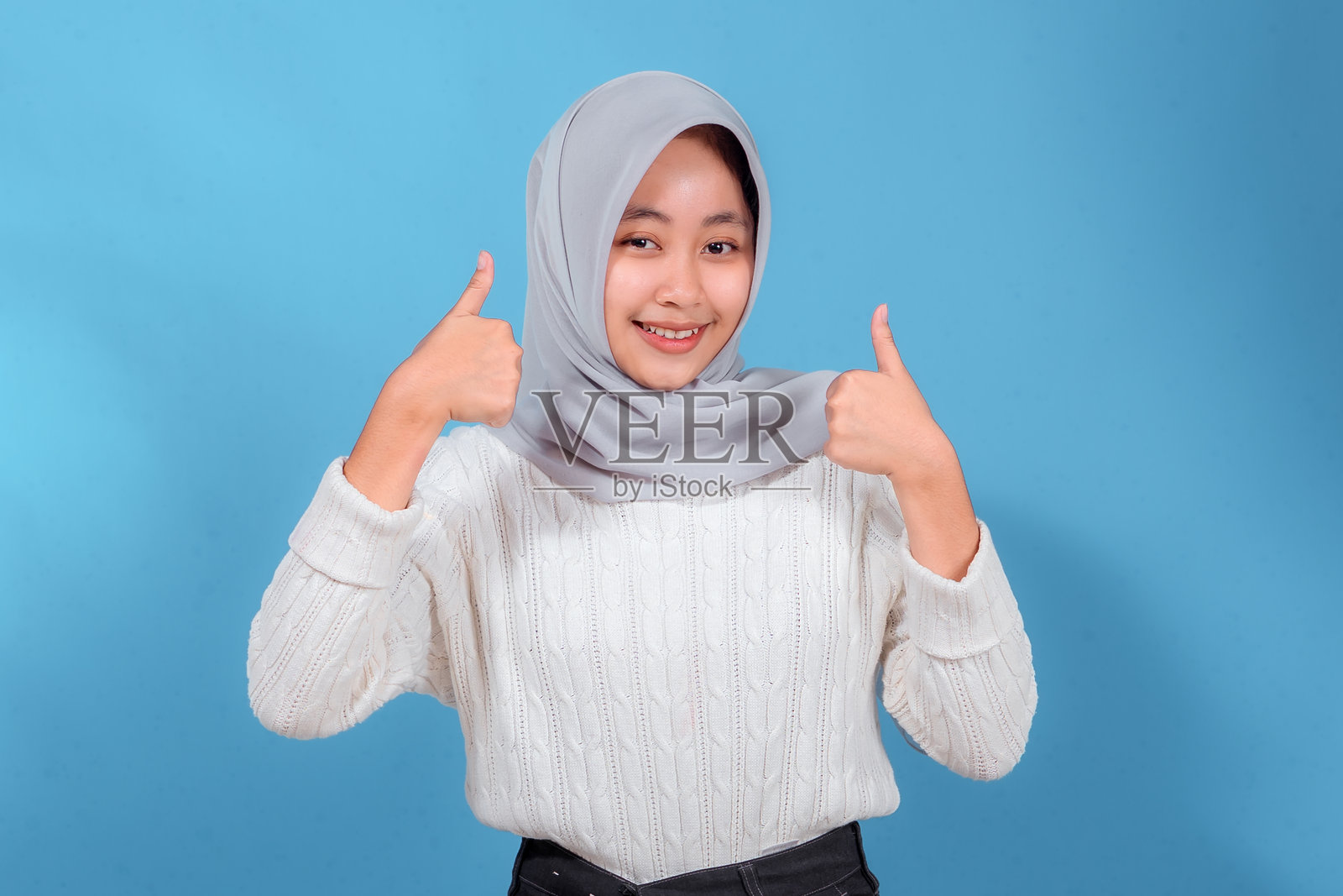 一位年轻女子戴着浅灰色头巾,身穿白色毛衣,在影棚中开心地举起双手点赞,脸上洋溢着自信的笑容,表达着喜悦、认可和成功。照片摄影图片