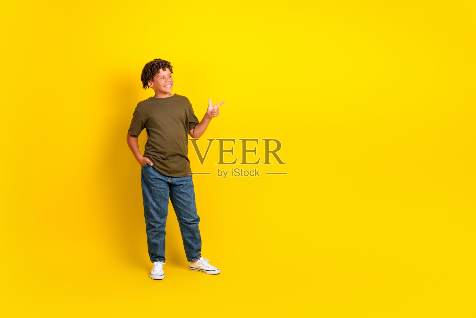 一个穿着休闲卡其色T恤、牛仔裤和运动鞋的有趣自信男孩,脸上带着微笑,指向前方,背景是明亮的黄色,营造出活泼的生活方式画面。照片摄影图片